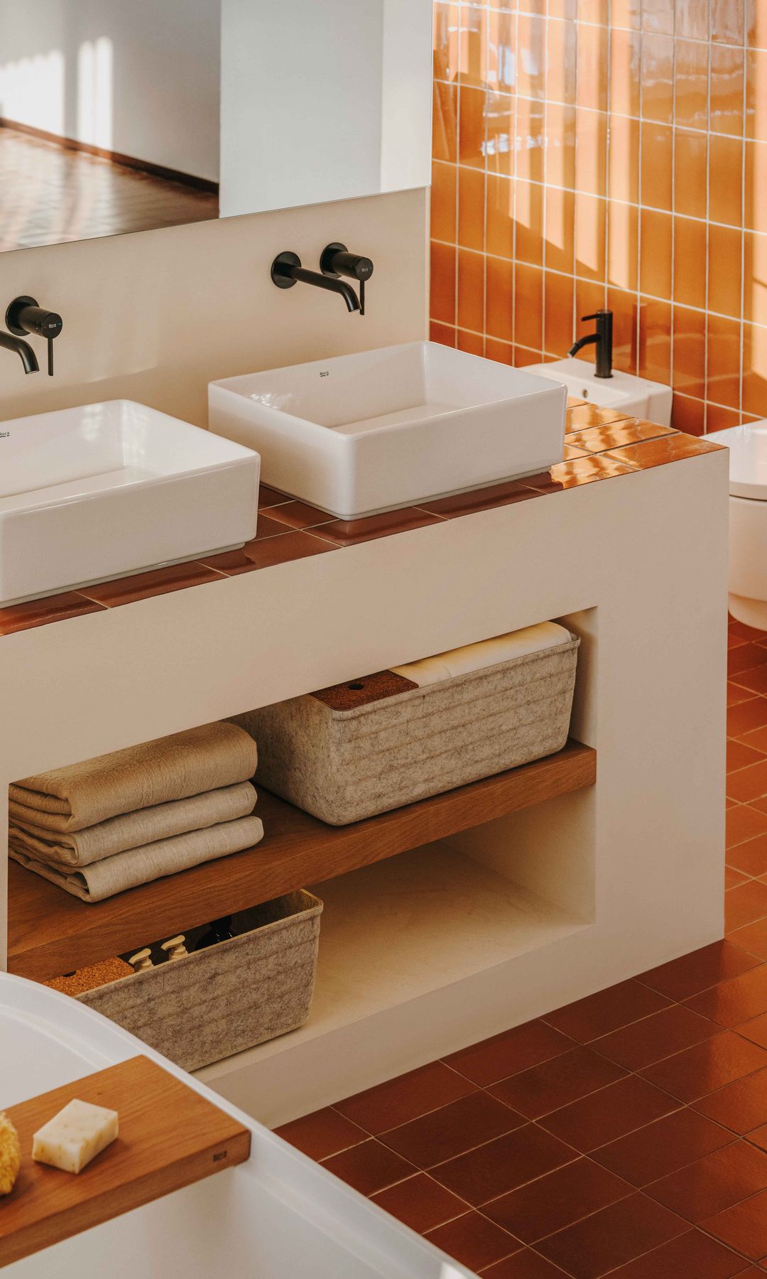 Baño con azulejos marrones, lavabos pequeños sobre encimera, griferías negras, bañera exenta, mueble bajolavabo de obra, balda de madera, cestos, toallas dobladas 