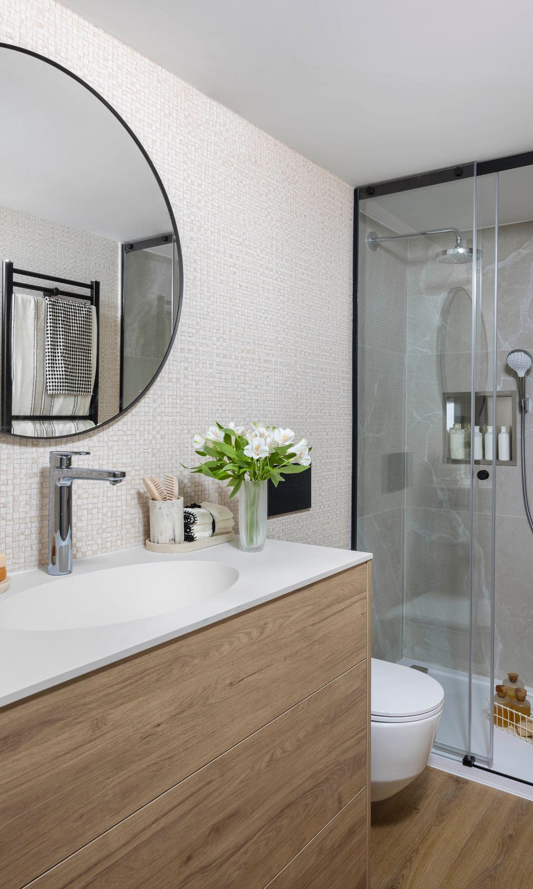 Baño con ducha y mampara con perfiles negros, mueble laminado con lavabo integrado, espejo redondo con el marco negro, suelo laminado efecto madera