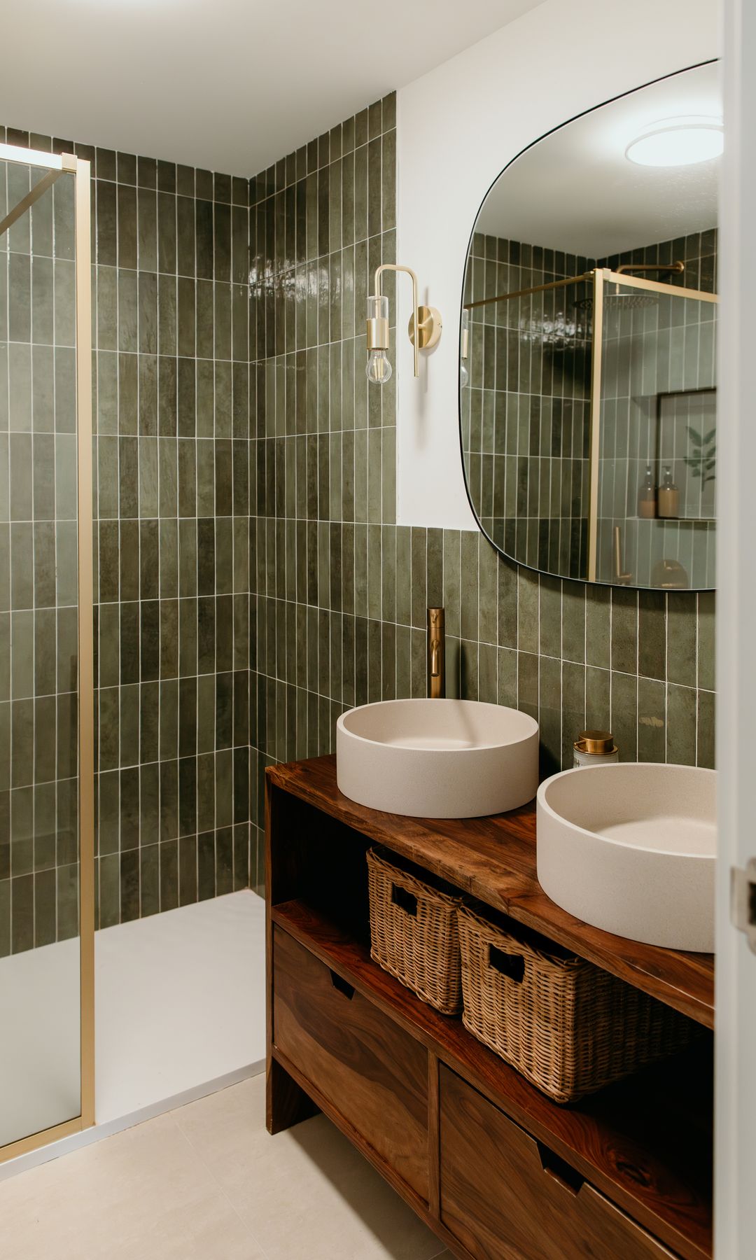Baño con ducha con mampara con perfiles dorados, mueble bajolavabo de madera, dos lavabos sobre encimera, espejo, cestas en el mueble, revestimiento de azulejos verdes