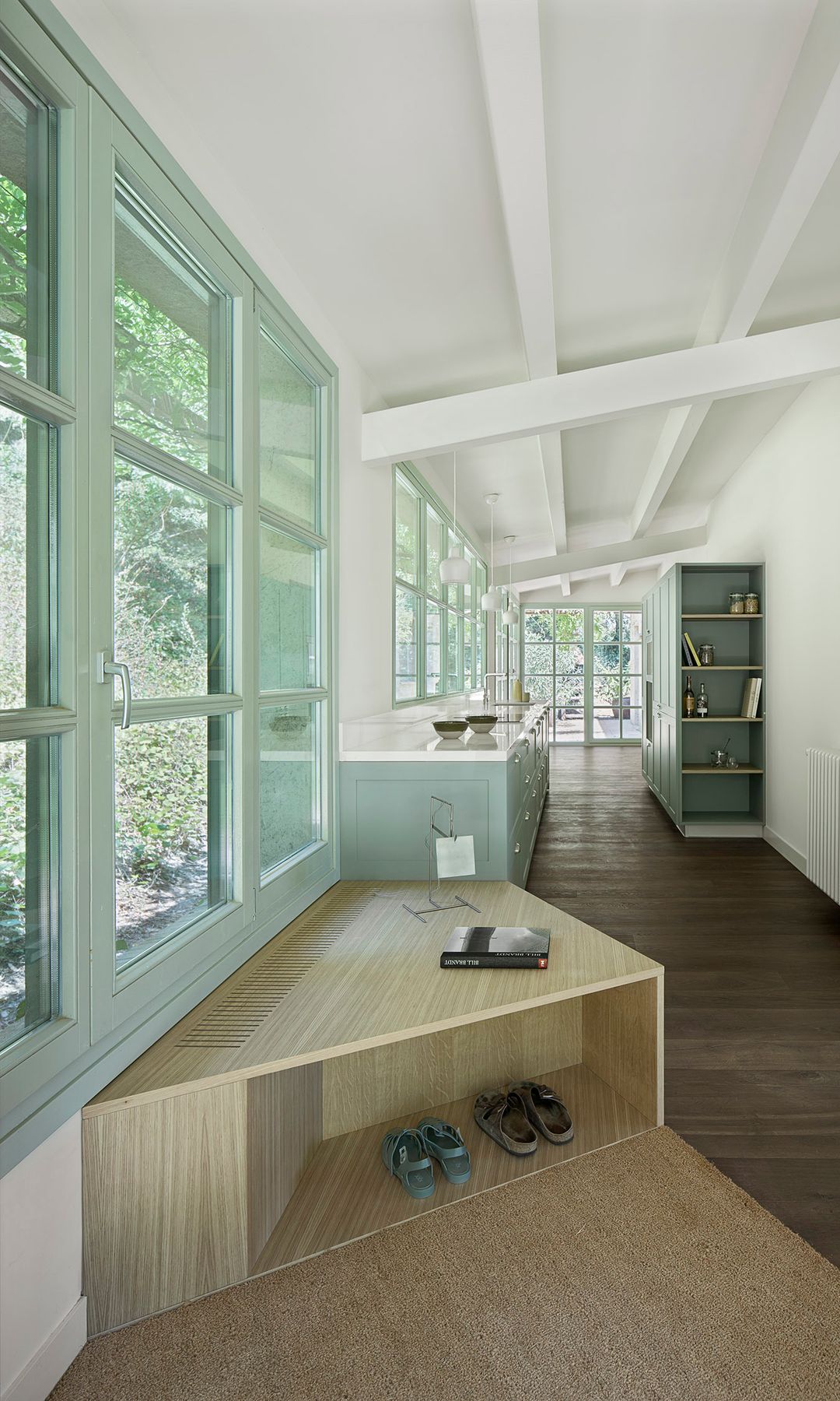 Recibidor abierto a la cocina con suelo de madera oscura, banco con zapatero integrado, ventanas con la carpintería de madera verde claro, muebles de cocina verde claro