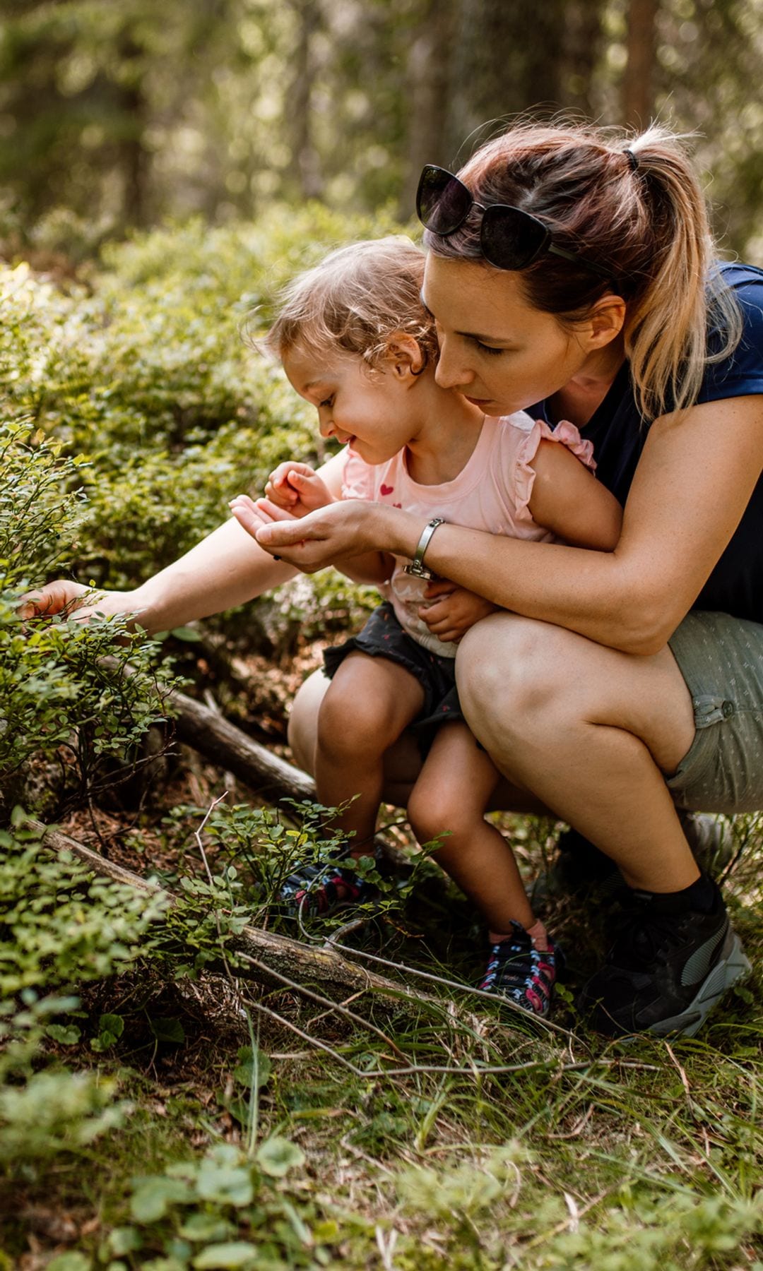 Madre con su hija cogiendo moras y plantas en el bosque, con ropa de verano, arrodilladas.