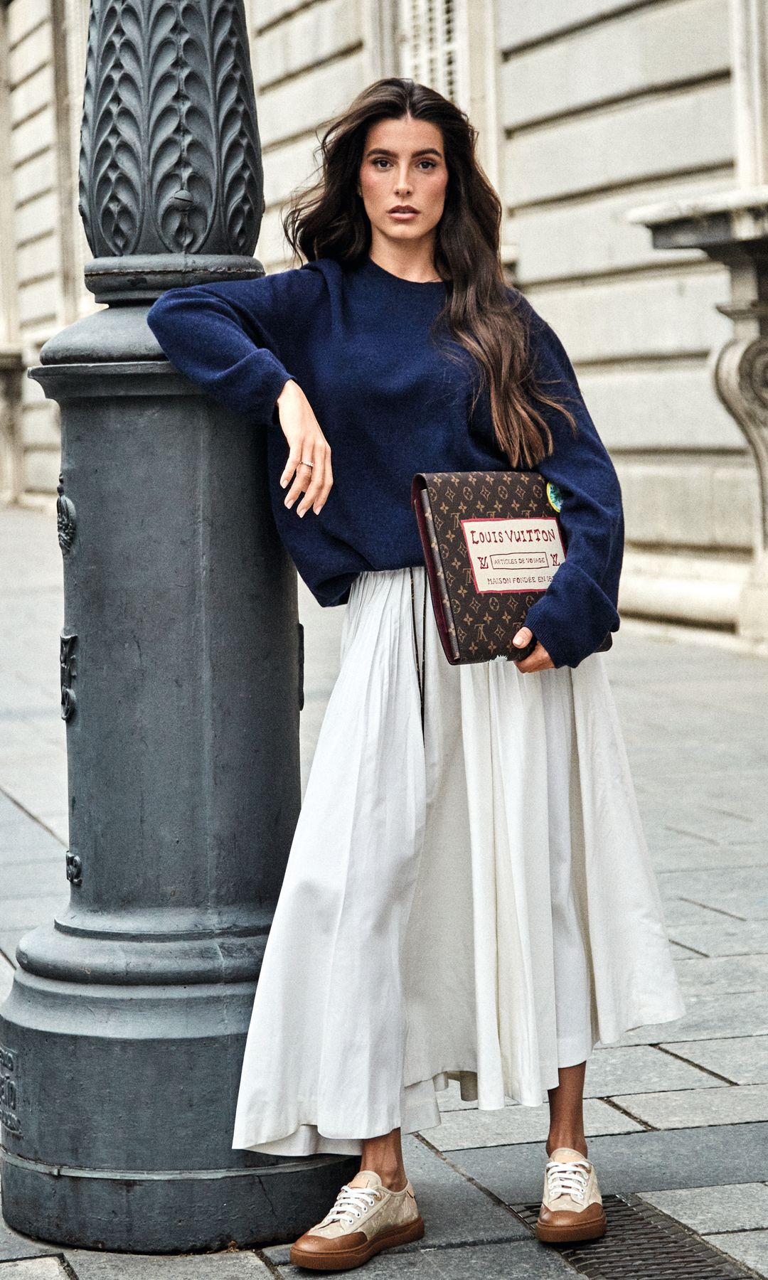 Aquí posa con jersey de cuello redondo, falda "midi" plisada, funda protectora en lona Monogram reinterpretada por Grace Coddington y zapatillas deportivas LV Lagoon, todo de Louis Vuitton