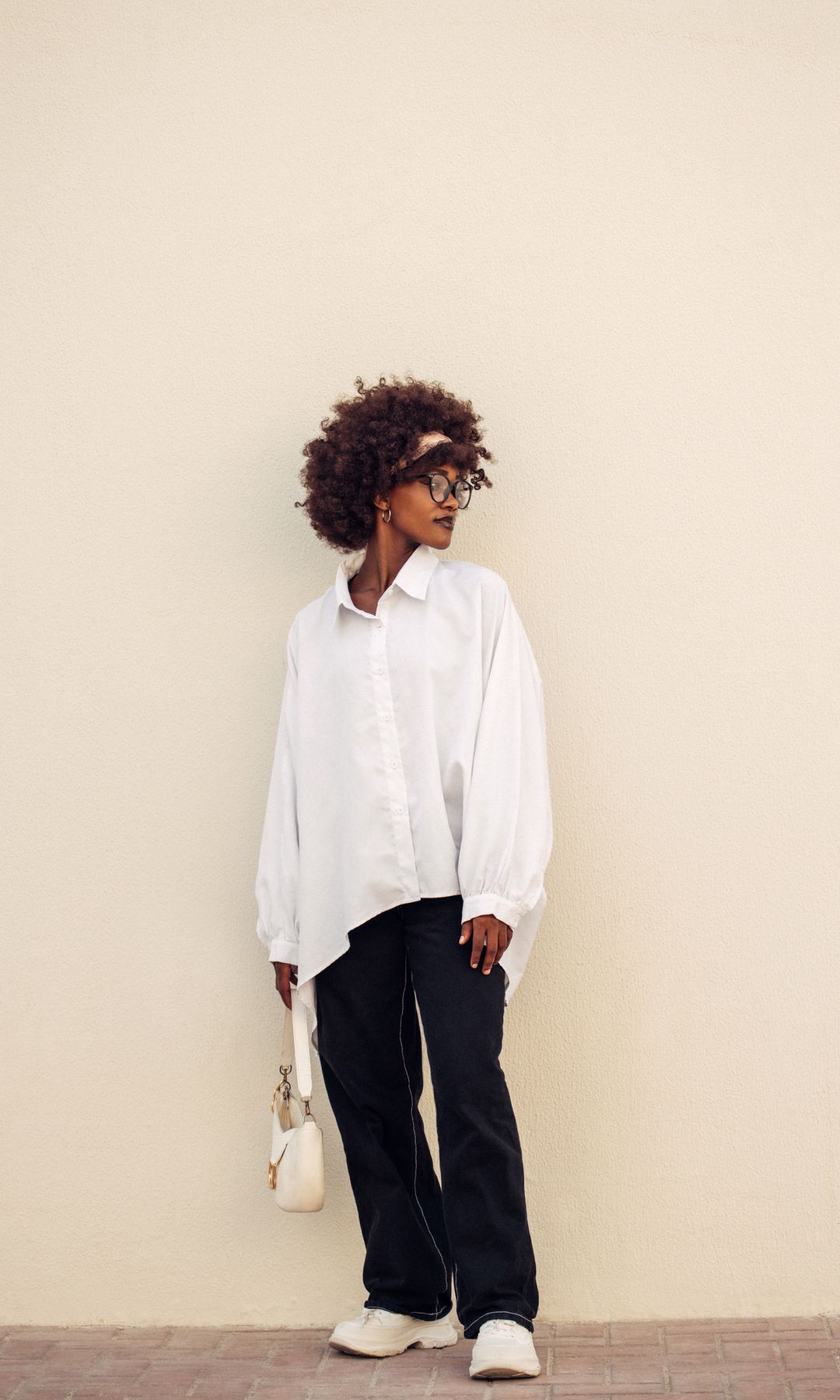 Mujer de color en una fotografía de cuerpo entero mirando hacia la izquierda, con camisa blanca oversize, pantalón negro y zapatillas deportivas blancas. Lleva gafas, un bolso en la mano en blanco.