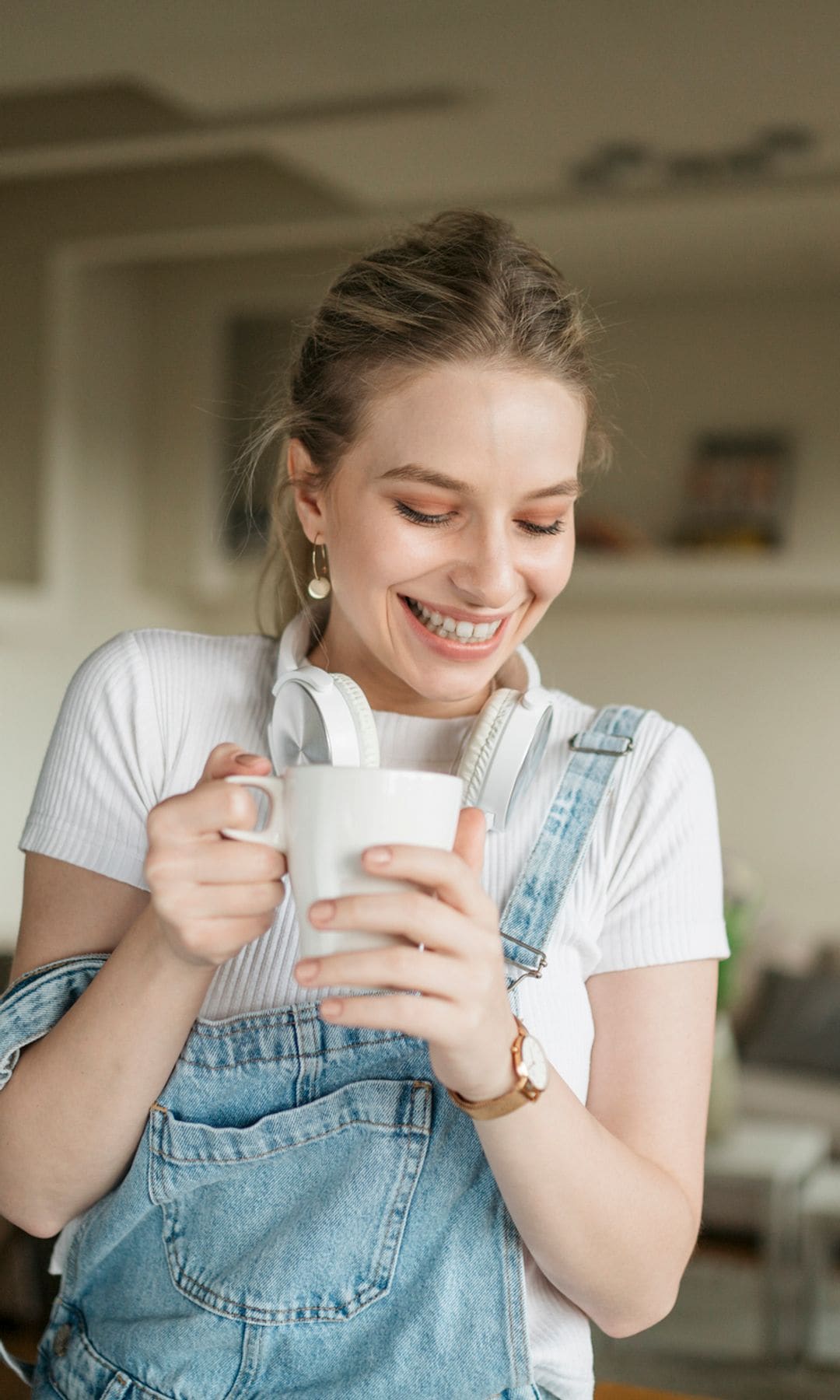 Mujer en overol azul sosteniendo una taza blanca, sonriendo en una sala de estar.