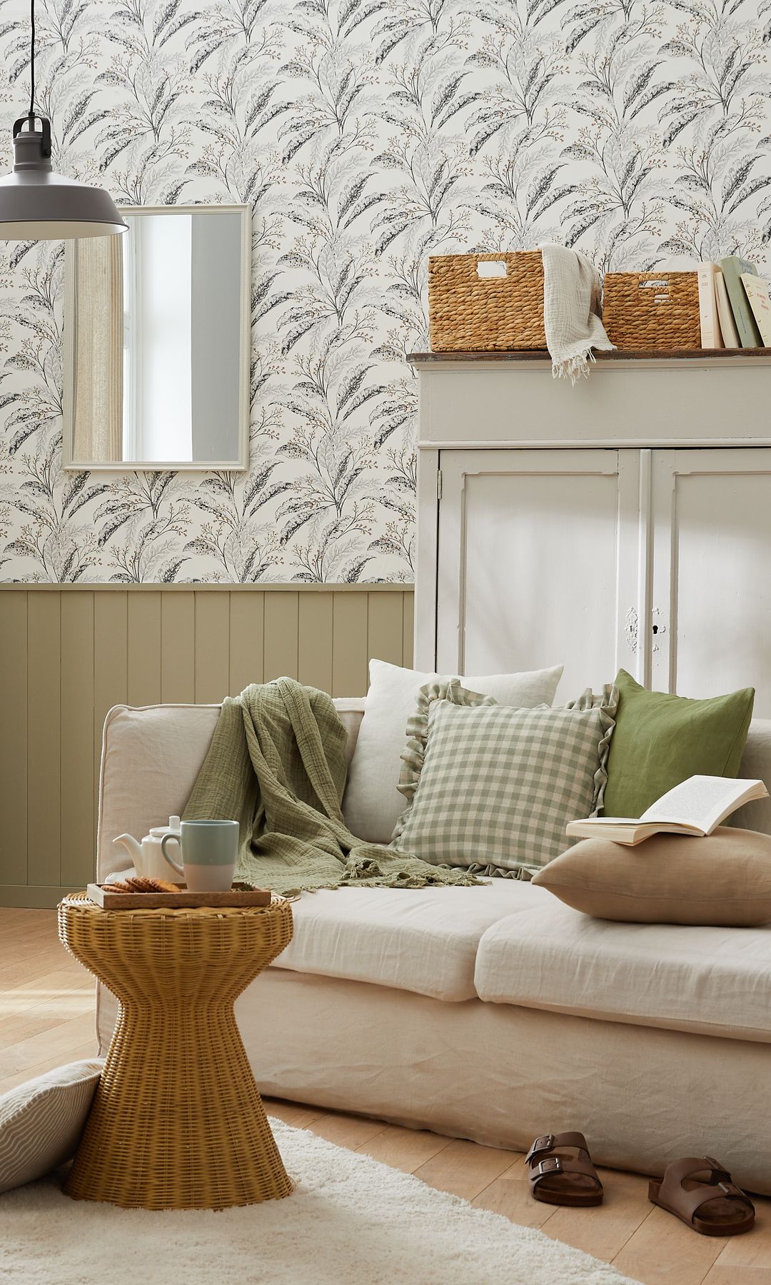 Salón con zócalo alto de madera verde, papel pintado de flores, sofá blanco, cojines lisos y de cuadros, manta verde, alfombra, mesa auxiliar de fibras vegetales 