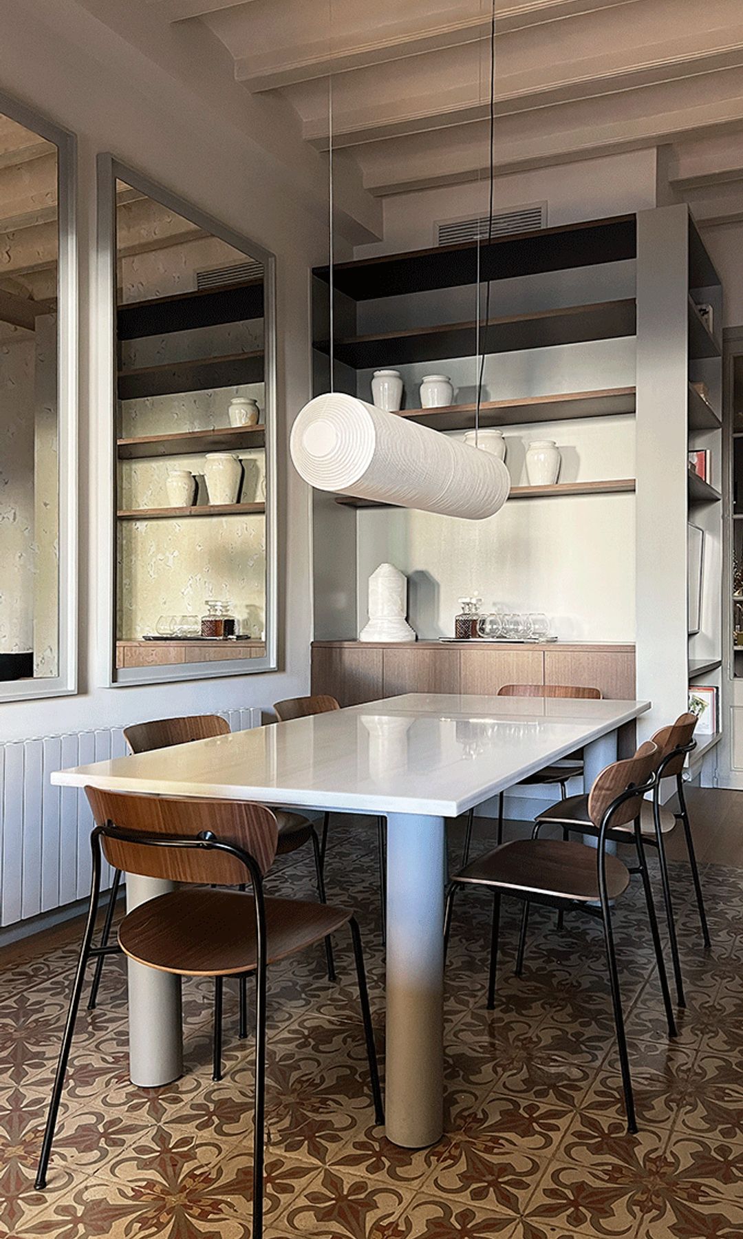 Comedor con mesa blanca, suelo hidráulico, sillas metálicas y con el asiento y el respaldo de madera oscura, baldas, aparador, lámpara de techo sobre la mesa