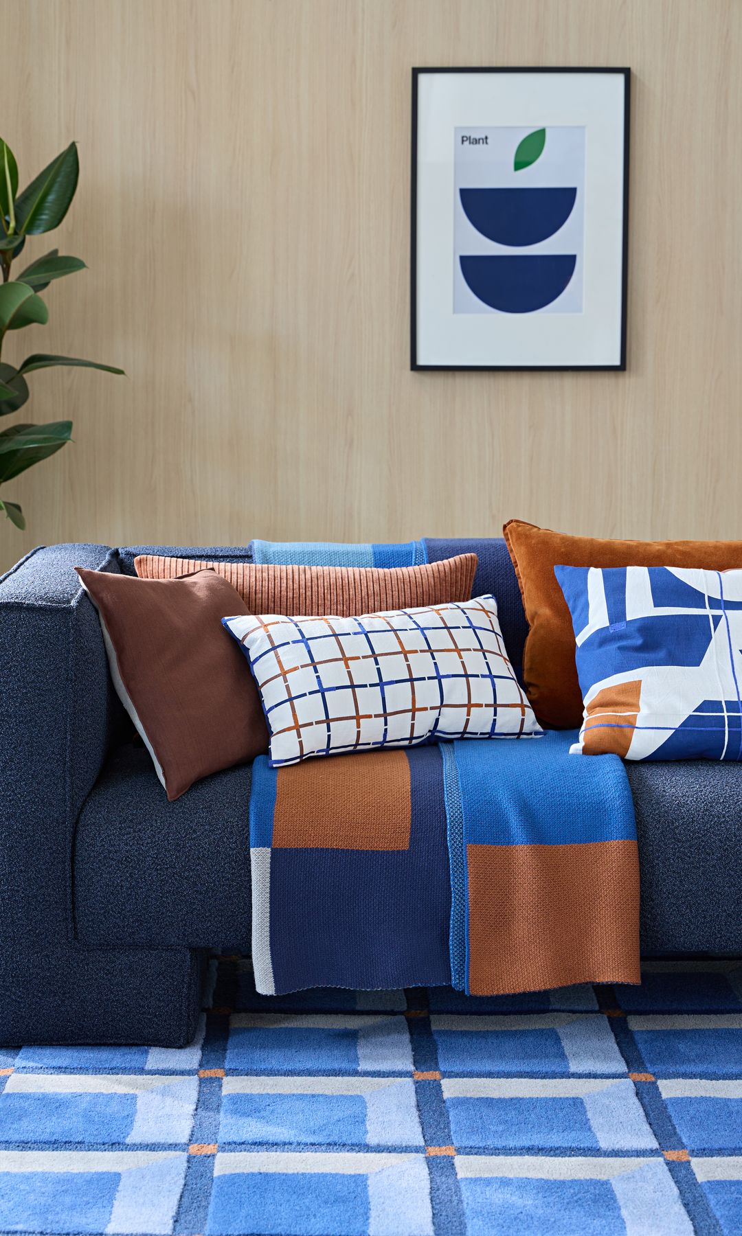 Salón con sofá en azul oscuro, alfombra en tonos azules, cojines marrones y azules, manta marrón y azul, cuadro en la pared, planta en maceta azul