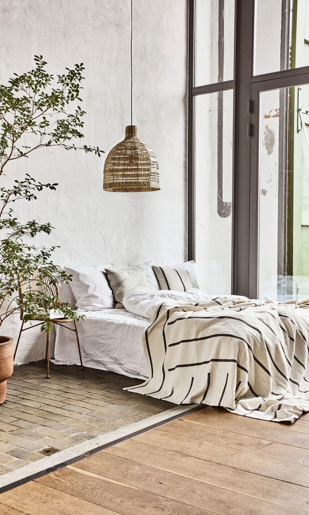 Dormitorio con lámpara de techo de fibras vegetales, gran planta verde, suelo de madera, ropa de cama de rayas blancas y negras