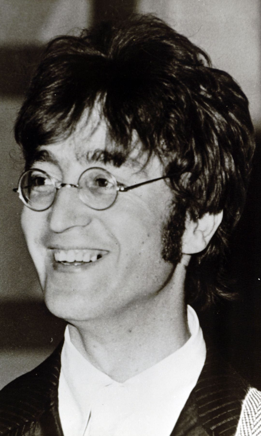 Foto en blanco y negro de John Lennon sonriendo, con lentes y traje.