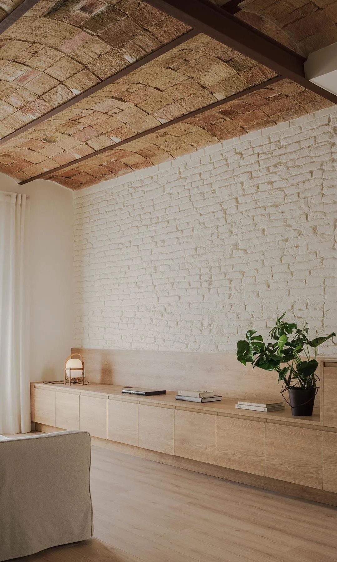 Salón con paredes de ladrillo pintadas de blanco, bóvedas catalanas en el techo, mueble de madera bajo frente el sofá, sofá blanco, suelo de madera, cortinas blancas