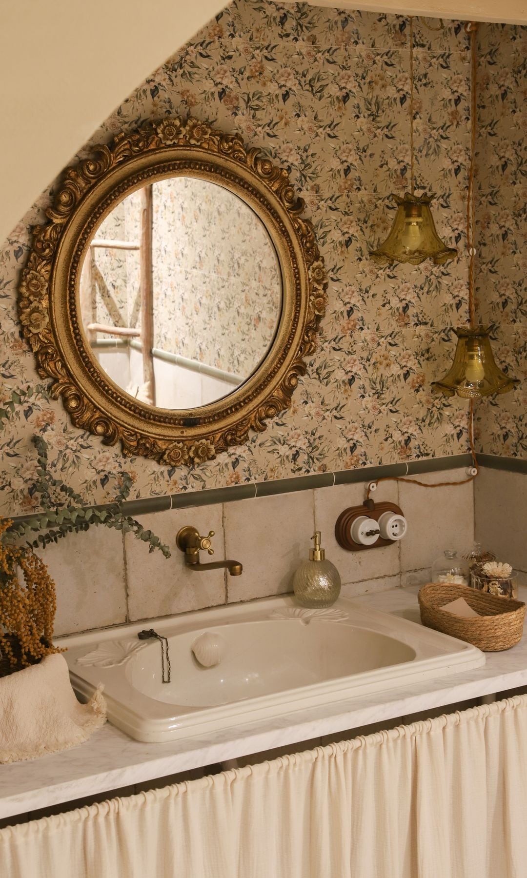 Baño antiguo con espejo redondo dorado, papel pintado de flores, lámparas de techo de cristal amarillo, llave de luz antigua, grifería dorada, mueble bajolavabo con cortina