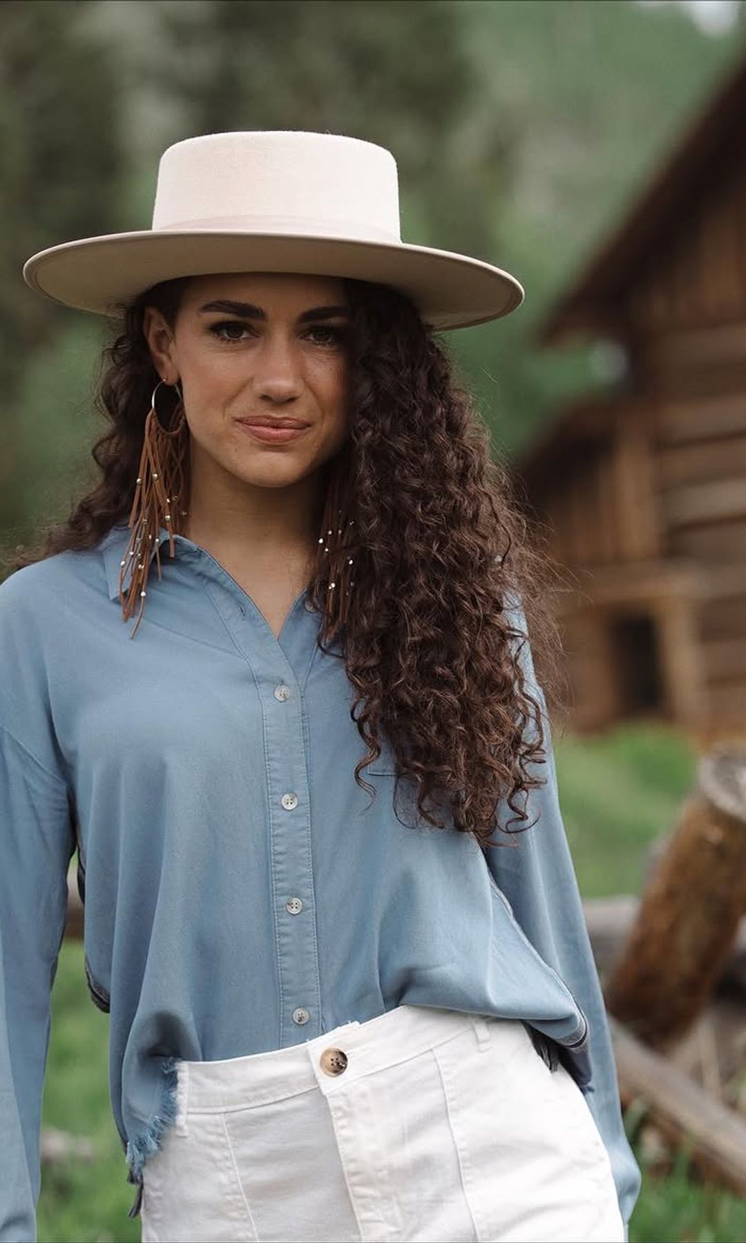 Mujer con larga melena castaña de rizos naturales definidos, sombrero de ala ancha y camisa azul en un entorno rural