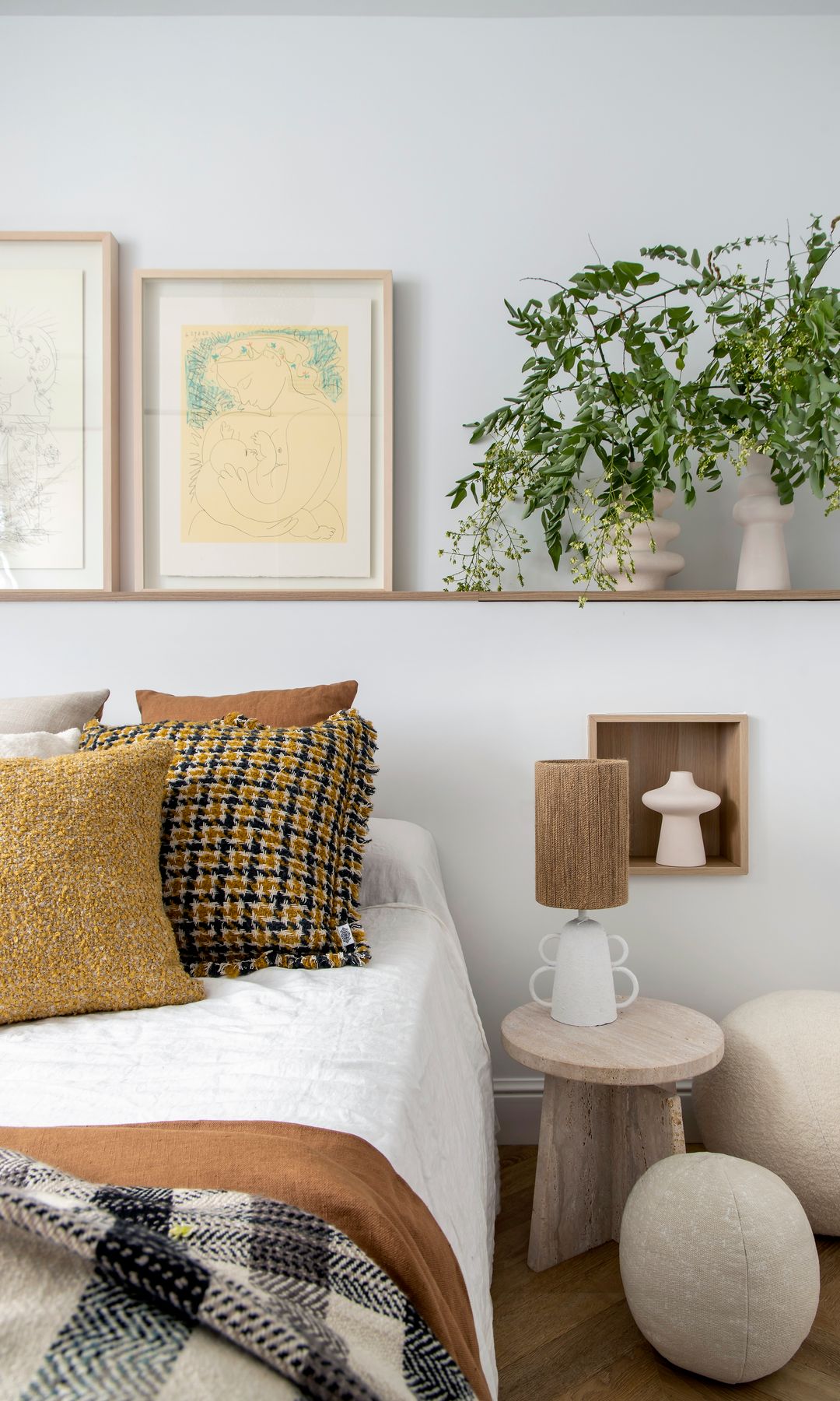 Dormitorio con cabecero de obra con calda de madera, jarrón con plantas, cuadros, cojines de color mostaza y cuadros, lámpara de sobremesa