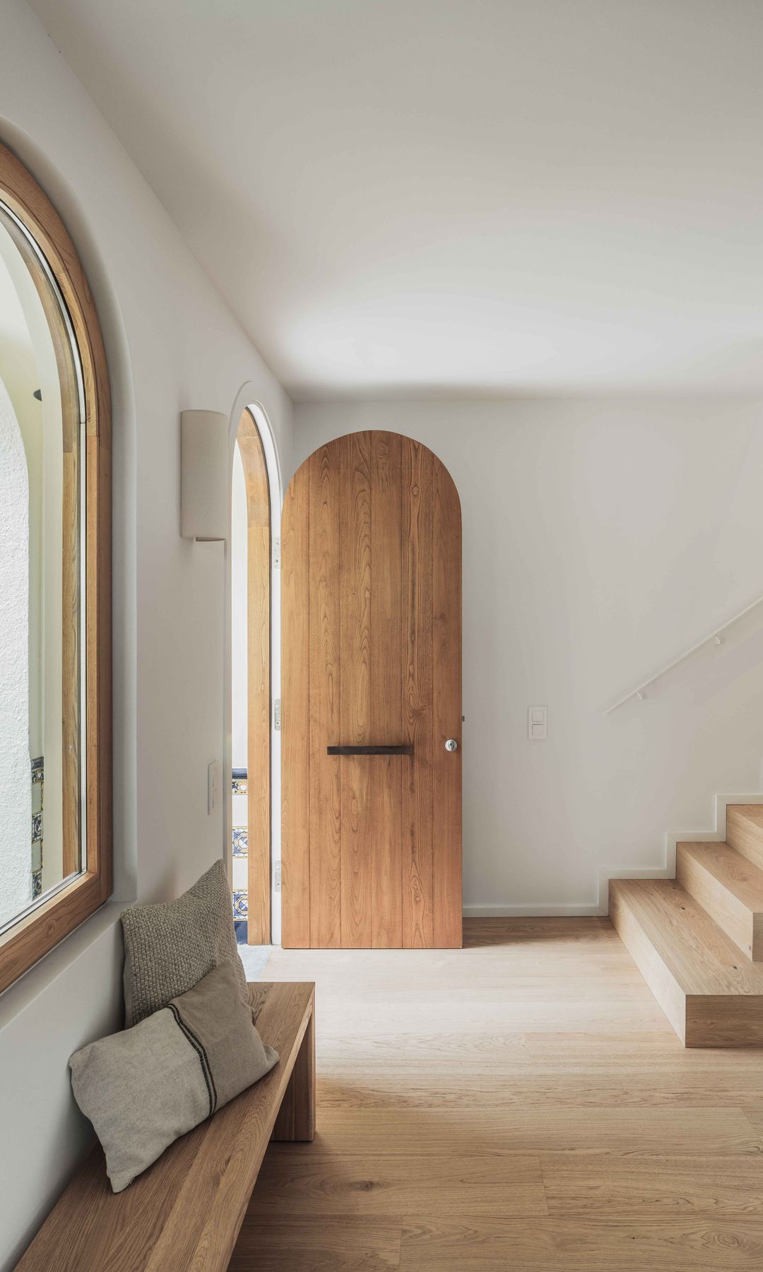 Recibidor con puerta de madera, banco de madera, escalera, armario blanco con hornacina de madera, ventana de medio punto de madera con vistas al jardín
