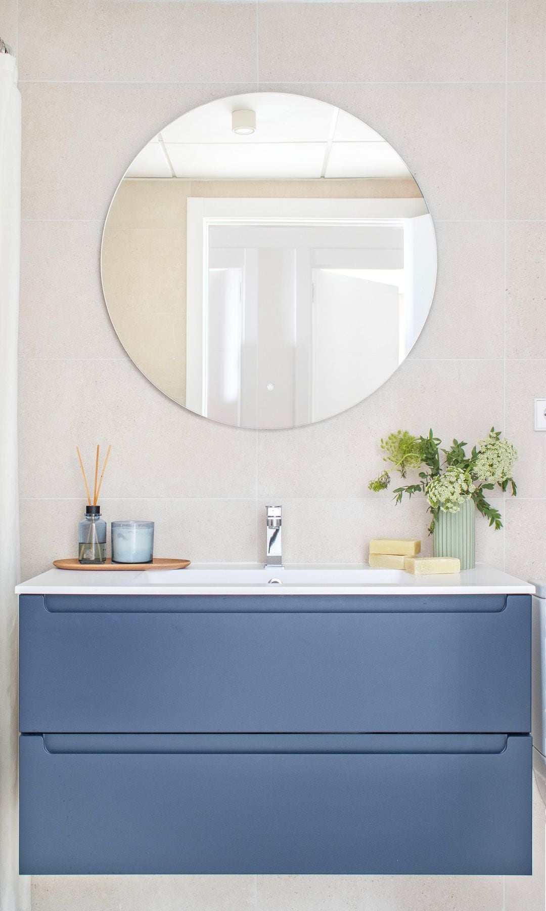 Baño con espejo redondo, mueble azul suspendido, jarrón con planta, bandeja con mikado