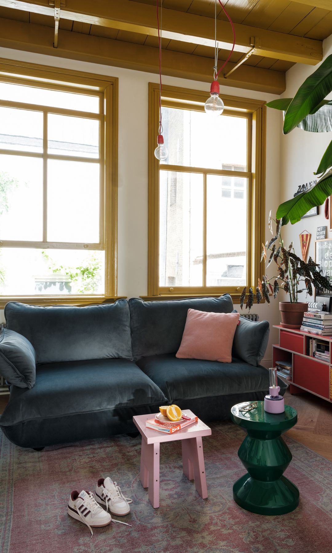 Salón con marcos de las ventanas amarillos, vigas metálicas en el techo amarillas, sofá de terciopelo azul oscuro, estantería baja roja, mesa auxiliar verde, taburete rosa, alfombra, cojín rosa