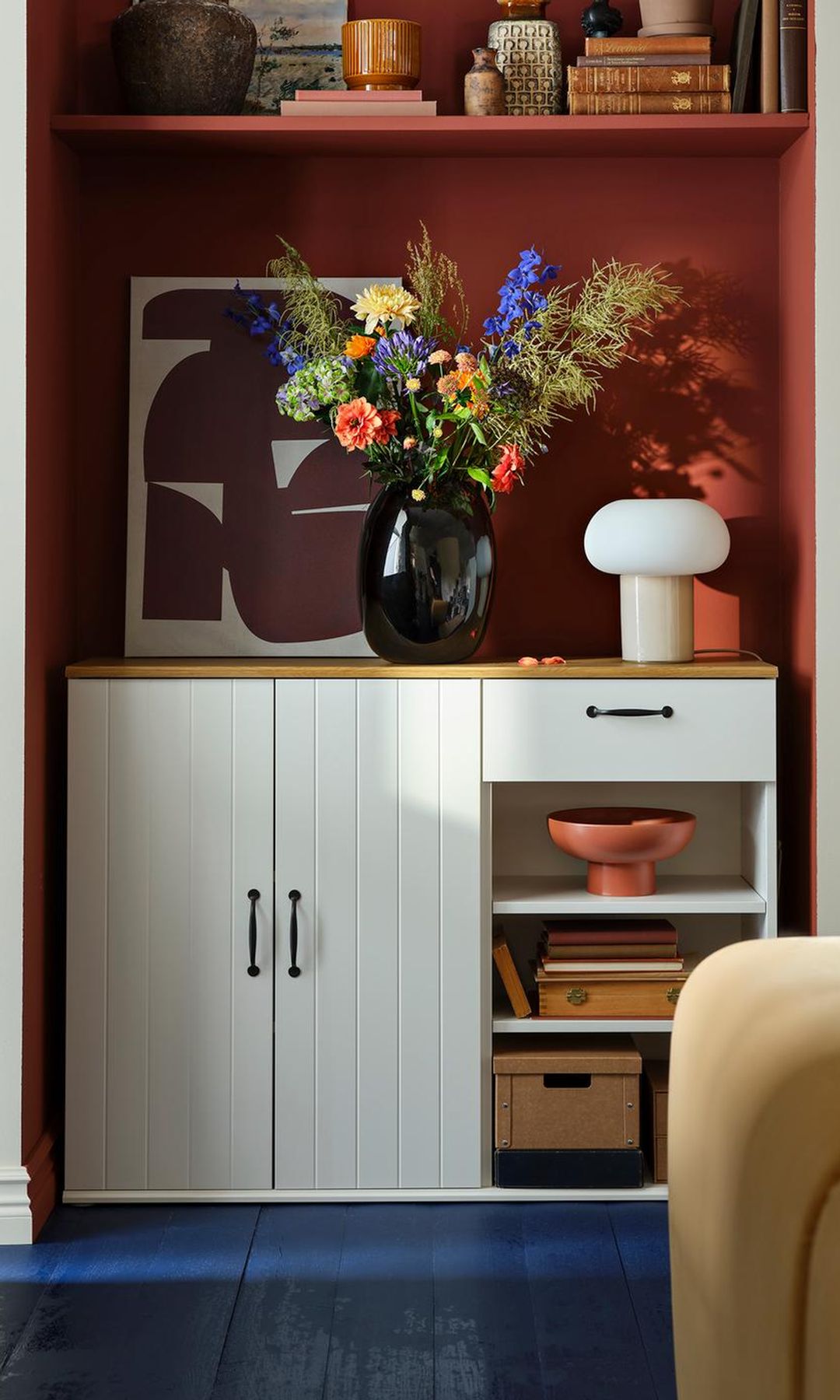 Aparador de IKEA en color blanco, con una parte con armarios y otra abierta con baldas y un cajón, pared de color marrón, jarrón negro con flores de colores, balda marrón, suelo azul 