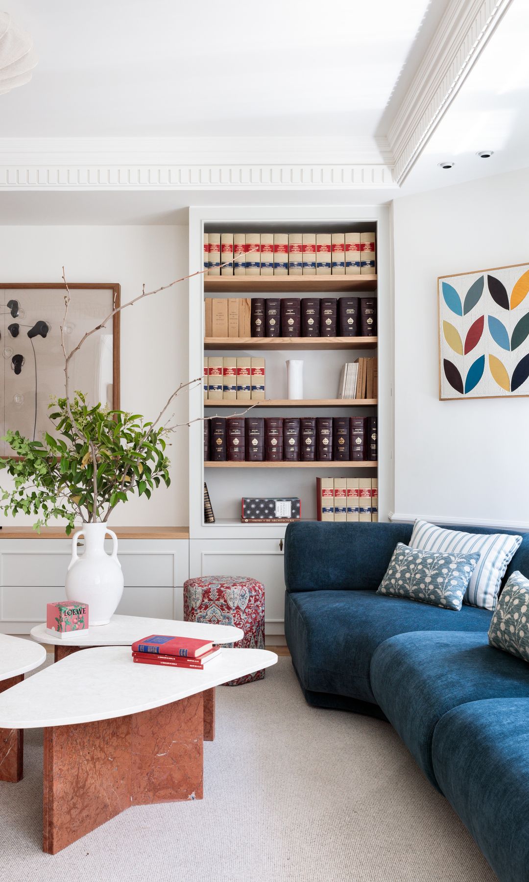 Salón con librería a medida de color blanco con. libros de derecho, sofá de color azul oscuro, cojines, mesas de centro con el sobre blanco, jarrón con plantas altas, cuadro en la pared 