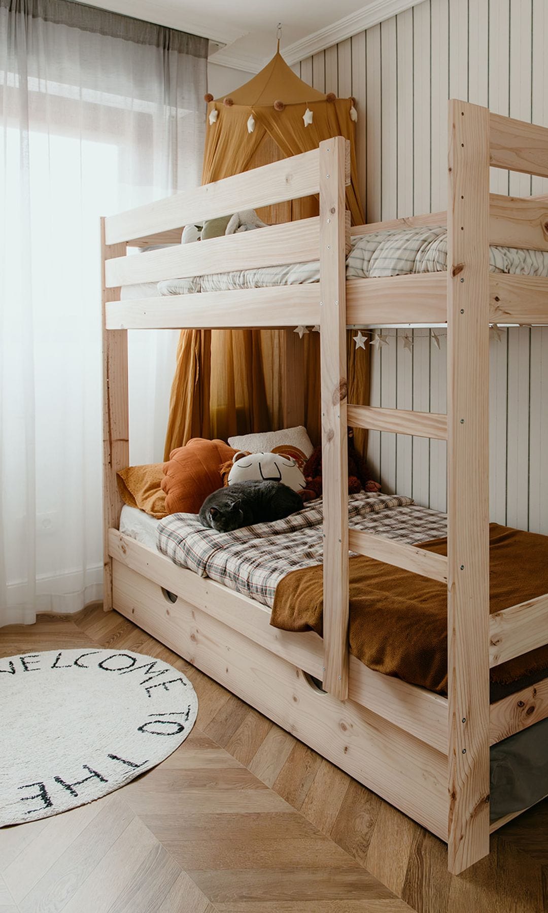 Habitación infantil con literas de madera, suelo de madera, alfombra redonda, cortinas blancas y papel pintado
