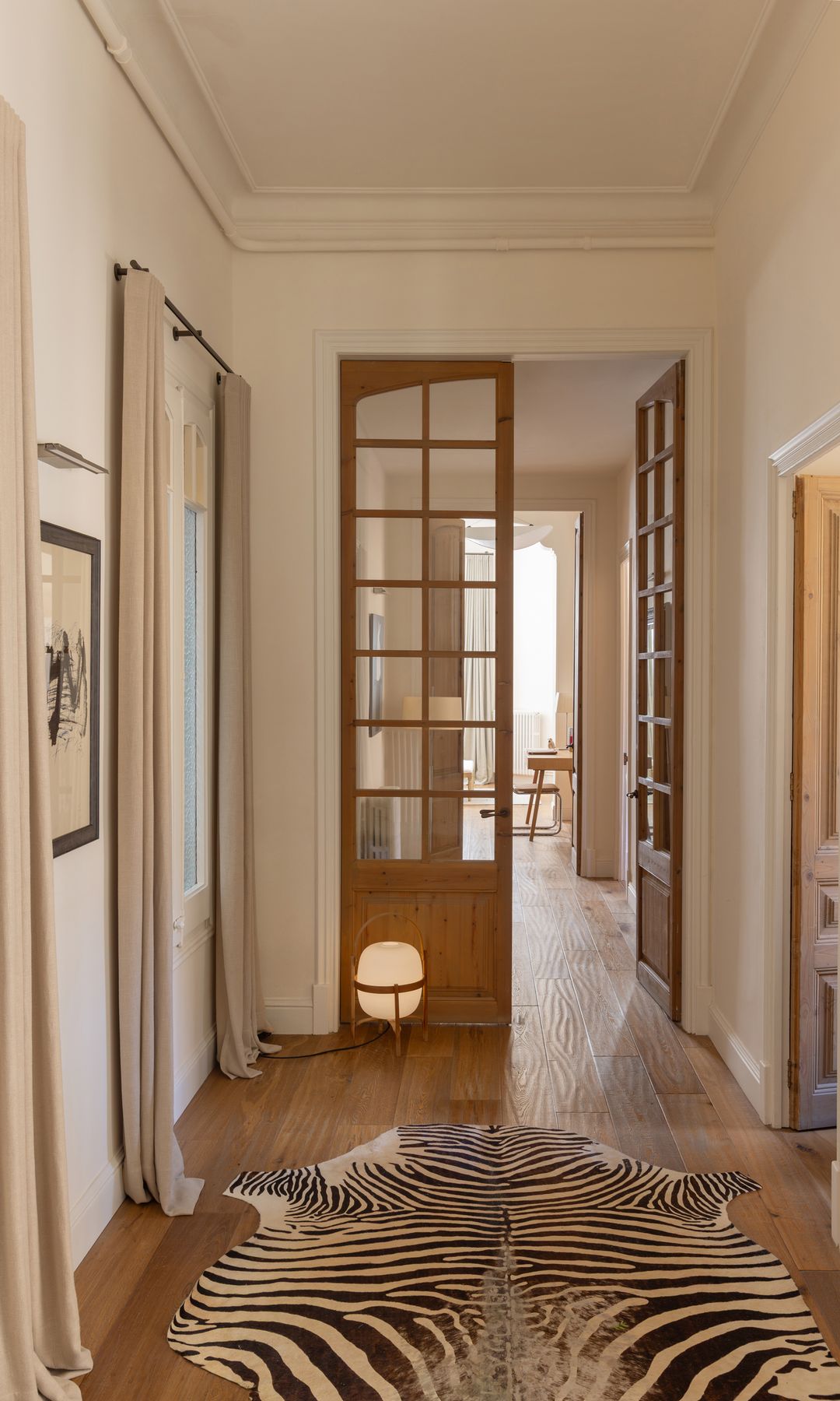 Pasillo distribuidor con puertas de madera con cuarterones de cristal, alfombra de piel de cebra, suelo de madera, lámpara de sobremesa Cesta, cortinas, cuadro en la pared con aplique 