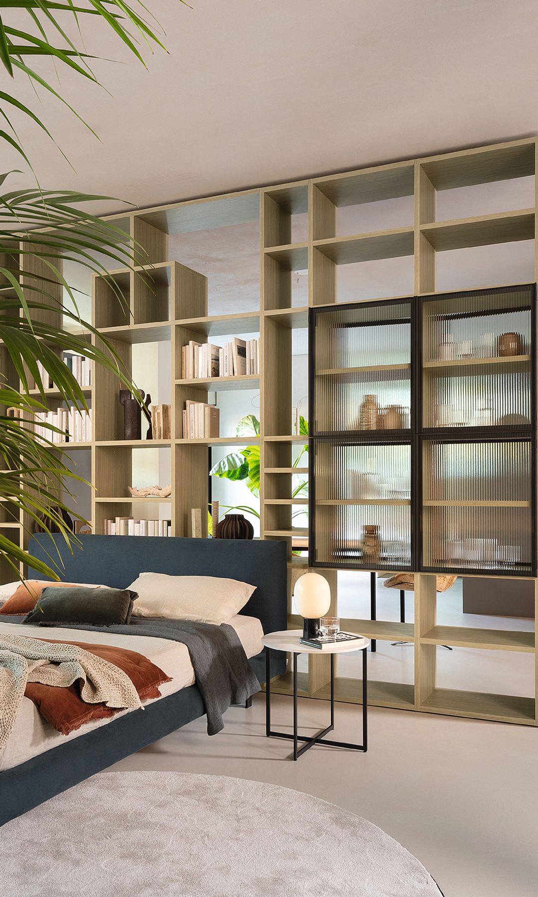 Dormitorio separado del salón por una librería sin trasera con una parte con vitrina, cama tapizada en azul 