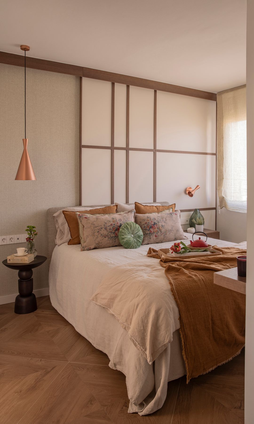 Dormitorio con pared del cabecero de aire japonés, cojines en colores tierra, suelo de madera, mesita de noche negra, lámpara de techo sobre la mesita 