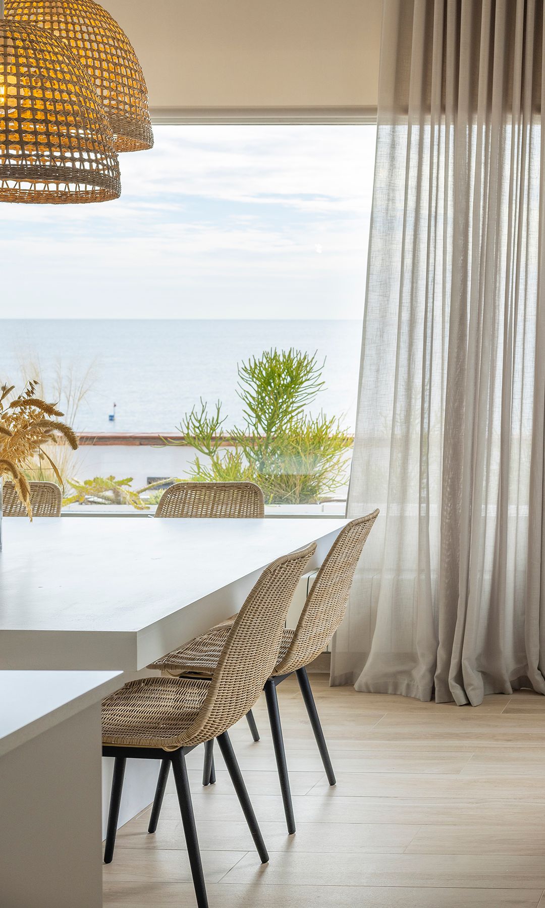 Comedor con gran ventanal, cortinas de lino, mesa blanca, sillas de fibras vegetales, suelo porcelánico con efecto madera, lámpara de techo de fibras vegetales
