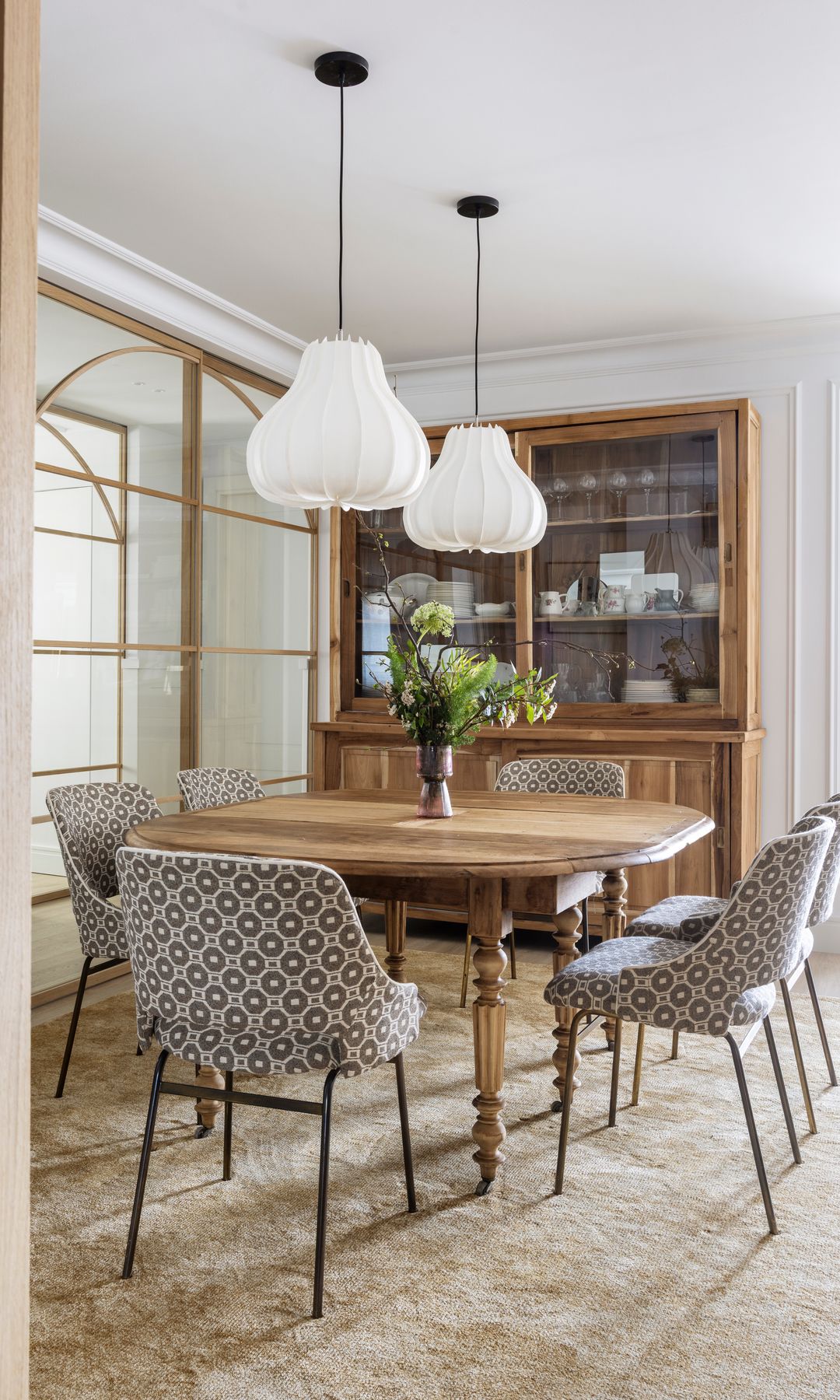 Comedor con mesa de madera con patas torneadas, alfombra, sillas tapizadas con tela motivos geométricos, vitrina, lámparas de techo blancas