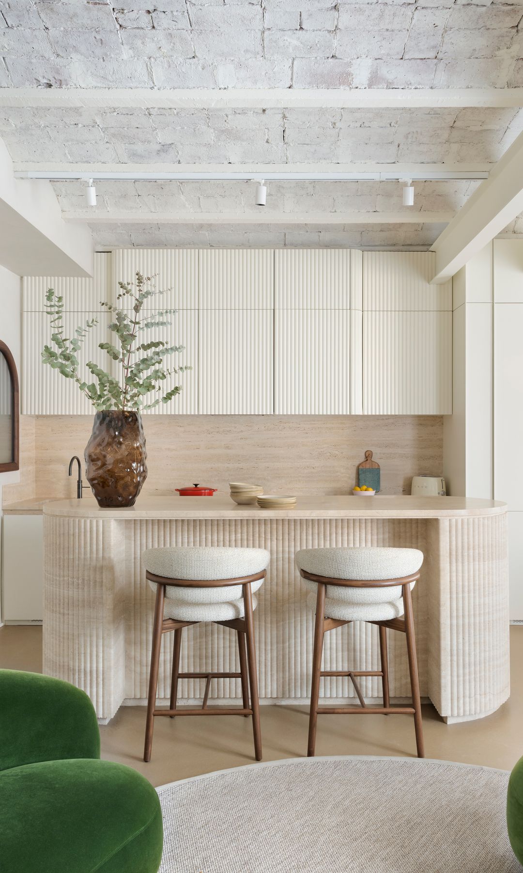 Cocina con isla con muebles beige, taburetes altos muebles hasta el techo, vigas y columnas, jarrón con rama verde sobre la encimera 
