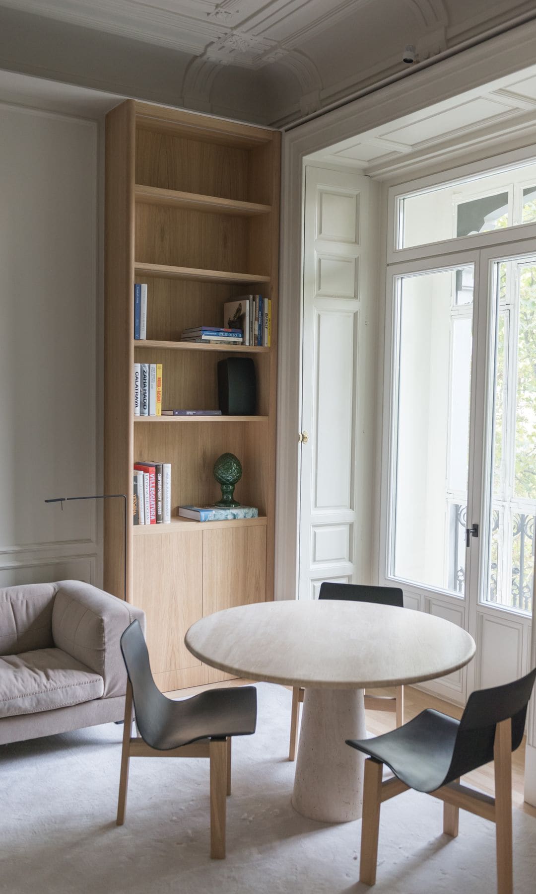Rincón del salón con mesa redonda, sillas, ventanal con contraventanas de madera blanca, estantería de madrea, alfombra, lámpara de pie 