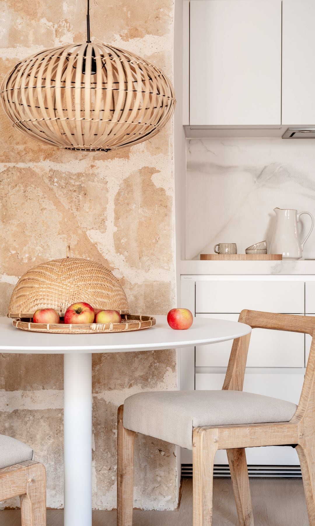 Comedor con cocina blanca, mesa redonda, sillas de madera tapizadas, lámpara de techo de fibras vegetales y frutero de fibras vegetales