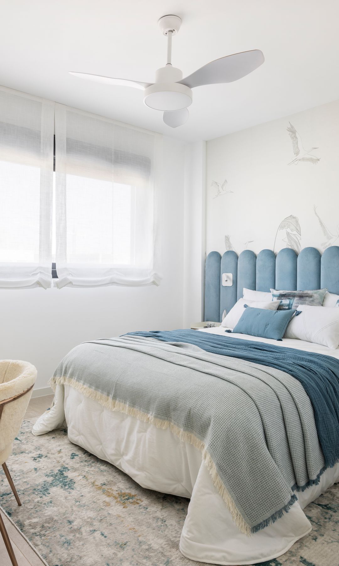 Dormitorio con ventilador de techo, cabecero tapizado en ondas azules, zona de escritorio, ventanas con estores blancos, ropa de cama azul, papel pintado en la pared del cabecero 