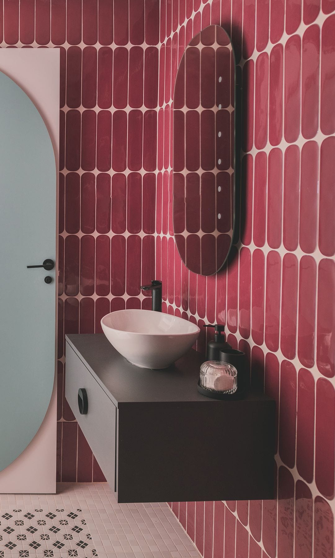 Baño pequeño con azulejos rojos, mueble con un cajón negro volado, lavabo sobre encimera, espejo, puerta con detalle en azul 
