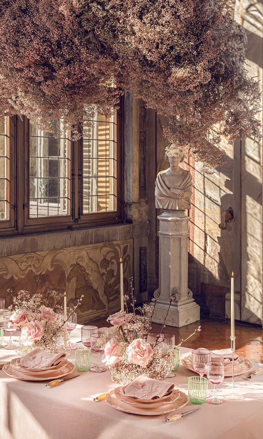 Comedor con mesa con mantel, vajilla en color rosa claro, copas en color rosa claro, arreglos florales en rosa, adorno colgante sobre la mesa en los mismos tonos, escultura clásica