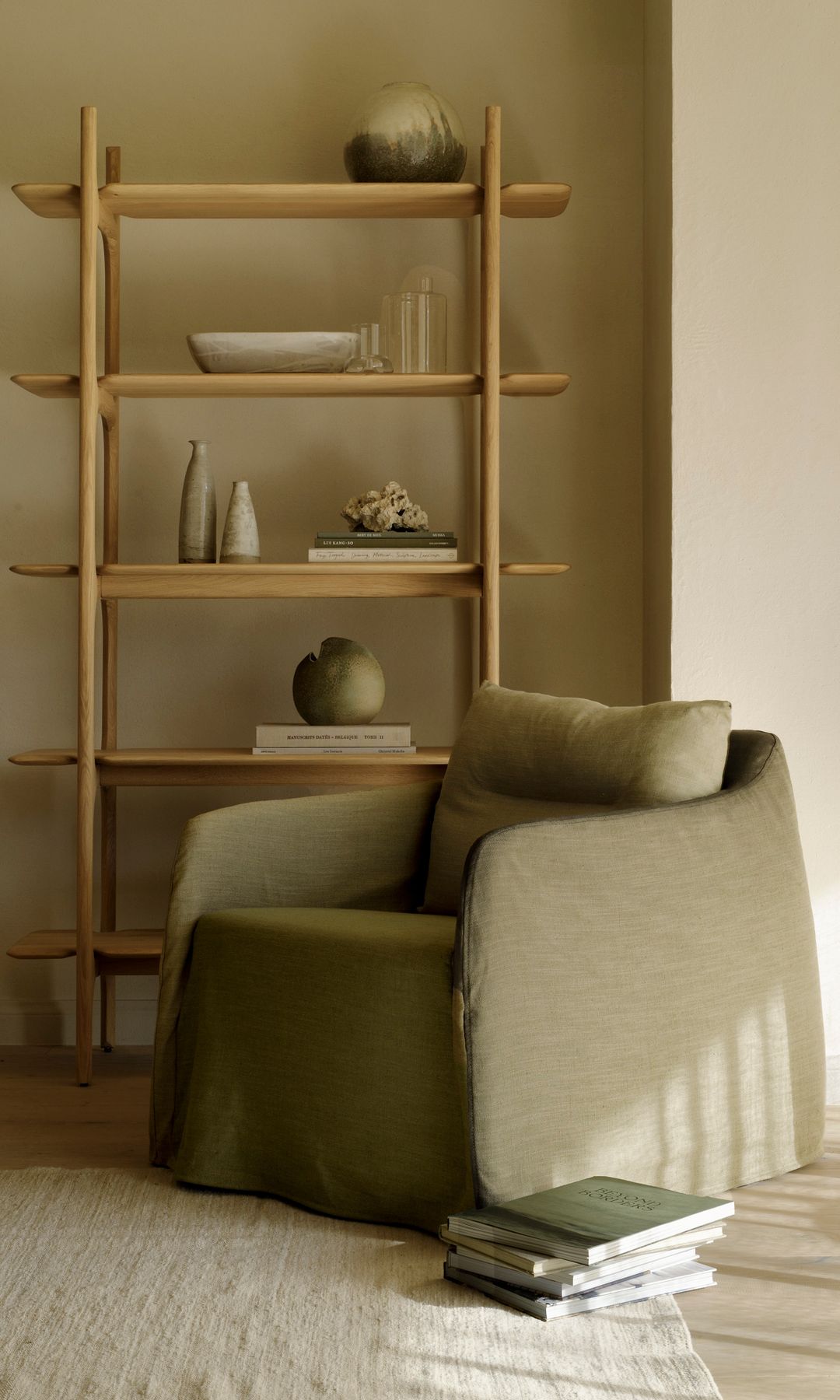 Salón con sillón verde oliva, estantería de madera sin trasera, alfombra, libros en el suelo 