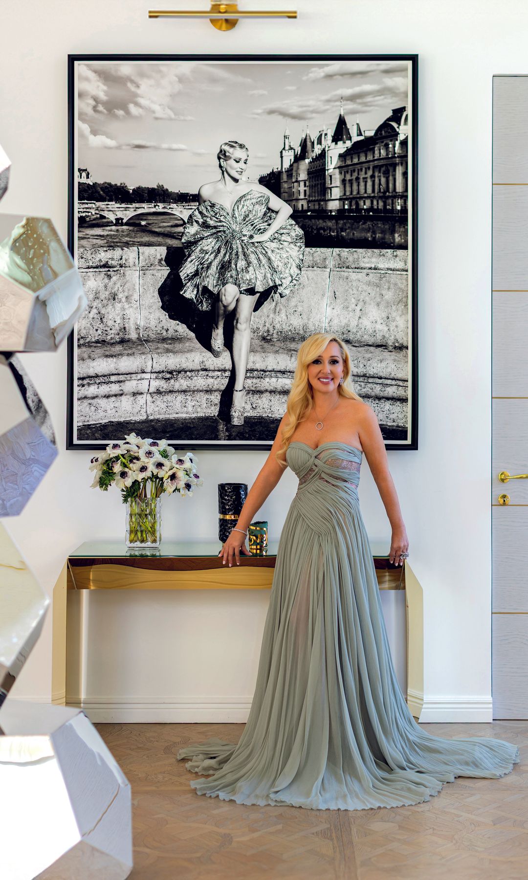 Mujer rubia en vestido plateado posando frente a una pintura grande.