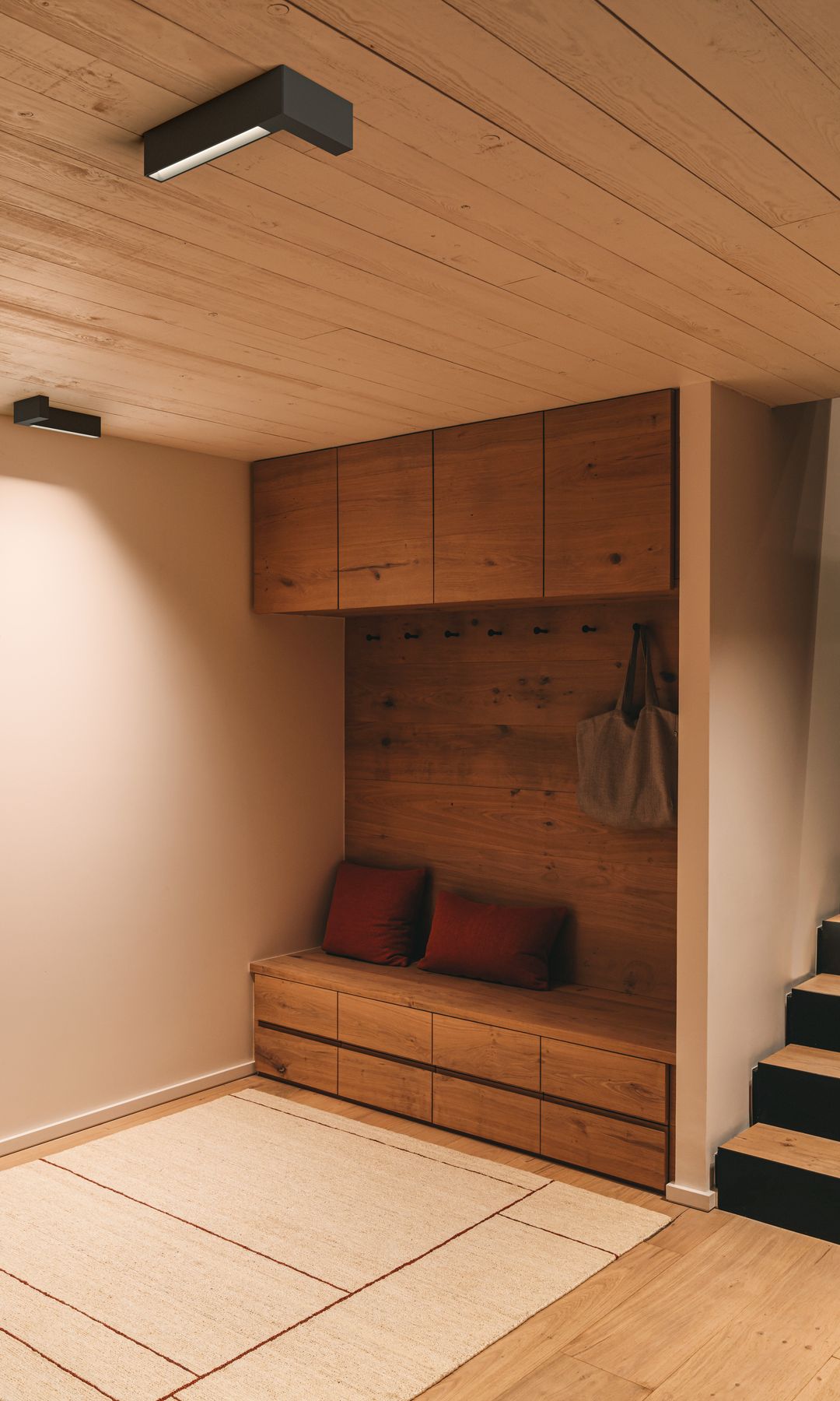 Sótano con techo de madera, lámpara de techo, alfombra, estructura de madera con ganchos para la ropa, banco, armarios superiores y cajones