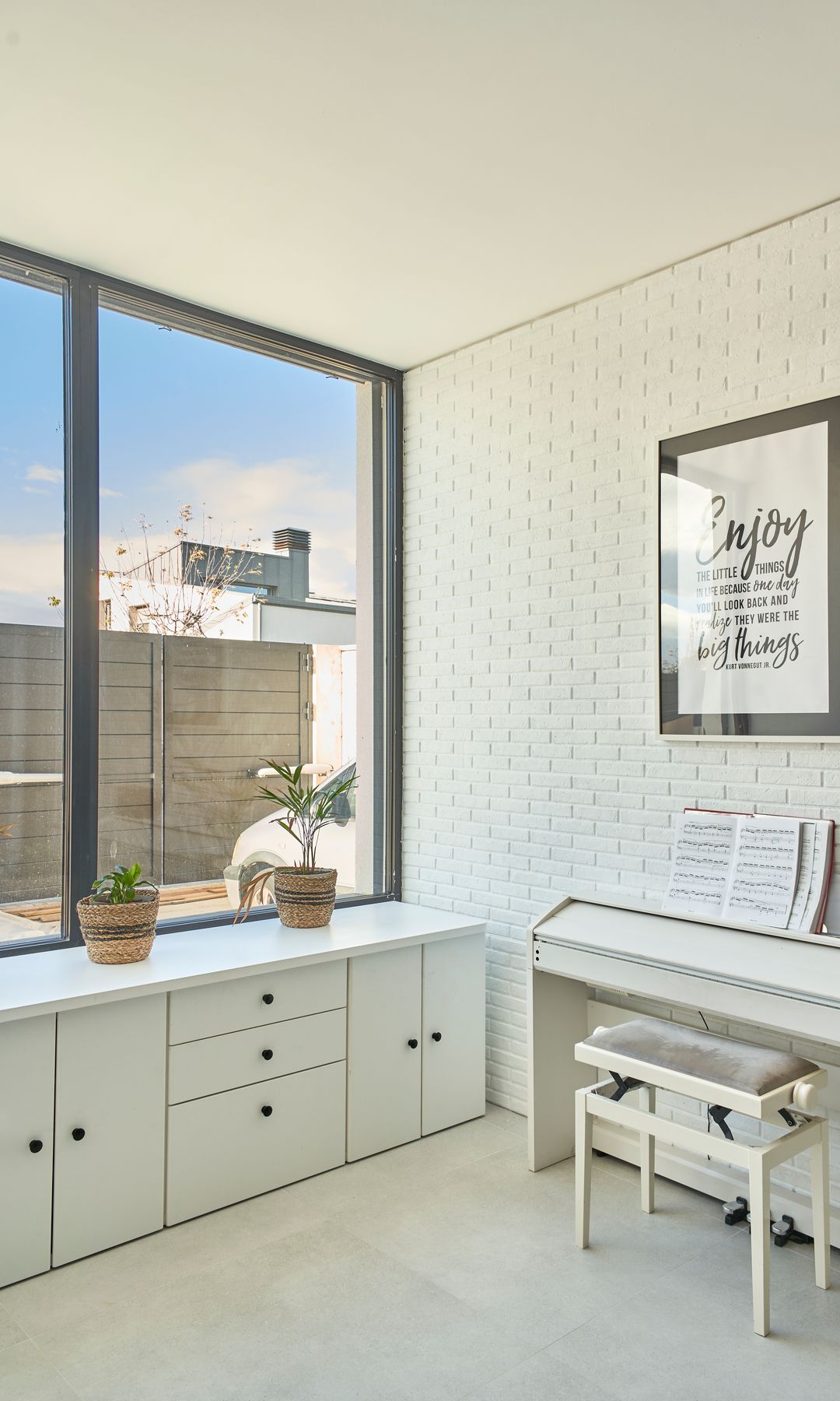 Habitación con pared de ladrillo blanco, gran ventanal. mueble bajo la ventana, piano blanco, cuadro en la pared
