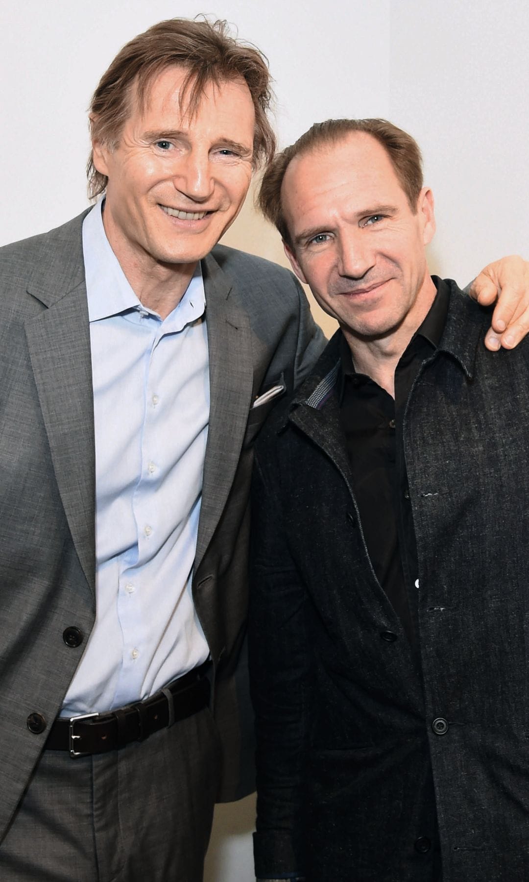 Liam Neeson y Ralph Fiennes sonrientes, Neeson en traje gris, Fiennes en chaqueta negra