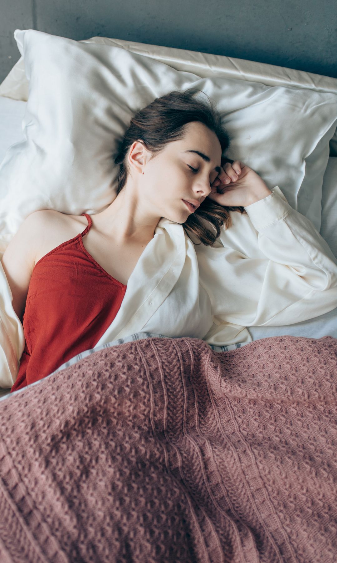 Mujer joven durmiendo sobre una almohada de seda, con camiseta roja y pijama de seda blanco. Está tapada a medio cuerpo con una manta rosa. Plano cenital.