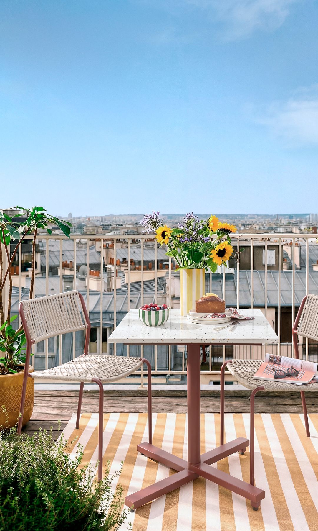 Terraza con mesa cuadrada pequeña, alfombra de rayas, sillas, macetas con plantas verdes, jarrón con tulipanes 