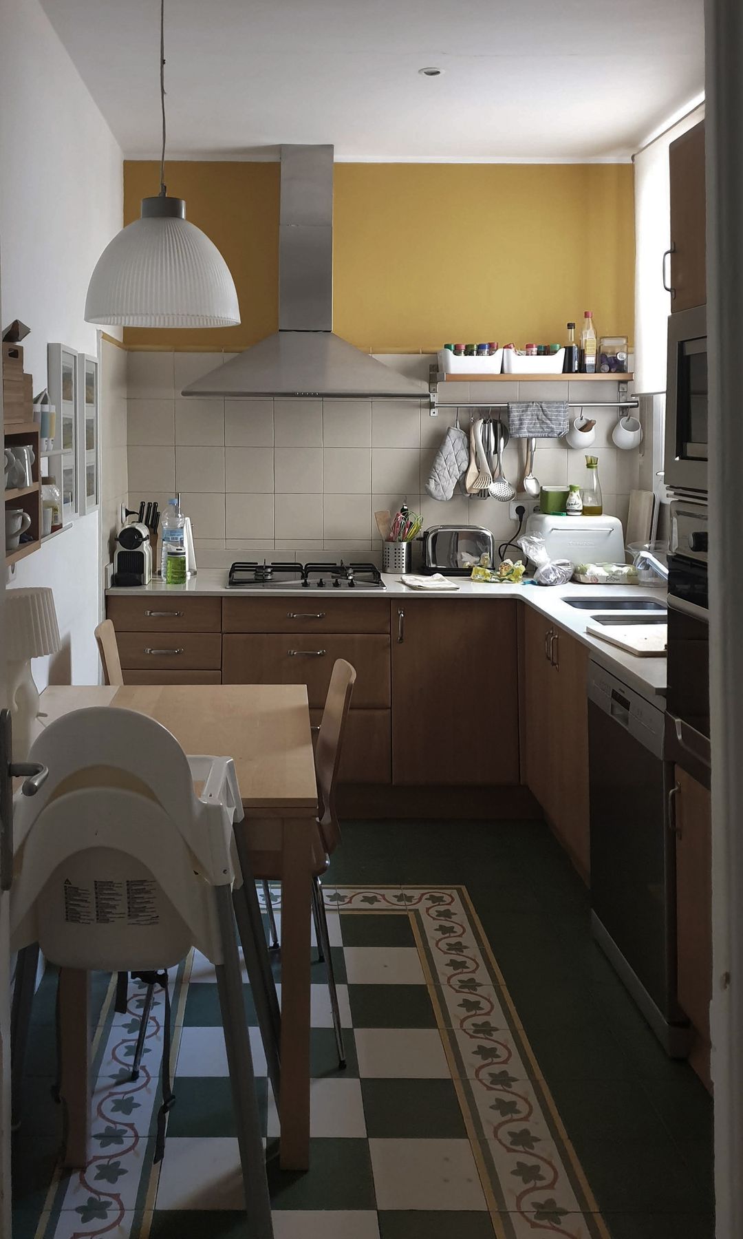 Cocina con suelo hidráulico, mesa de comedor, lámpara de techo, muebles de madera y armarios superiores amarillos, trona infantil