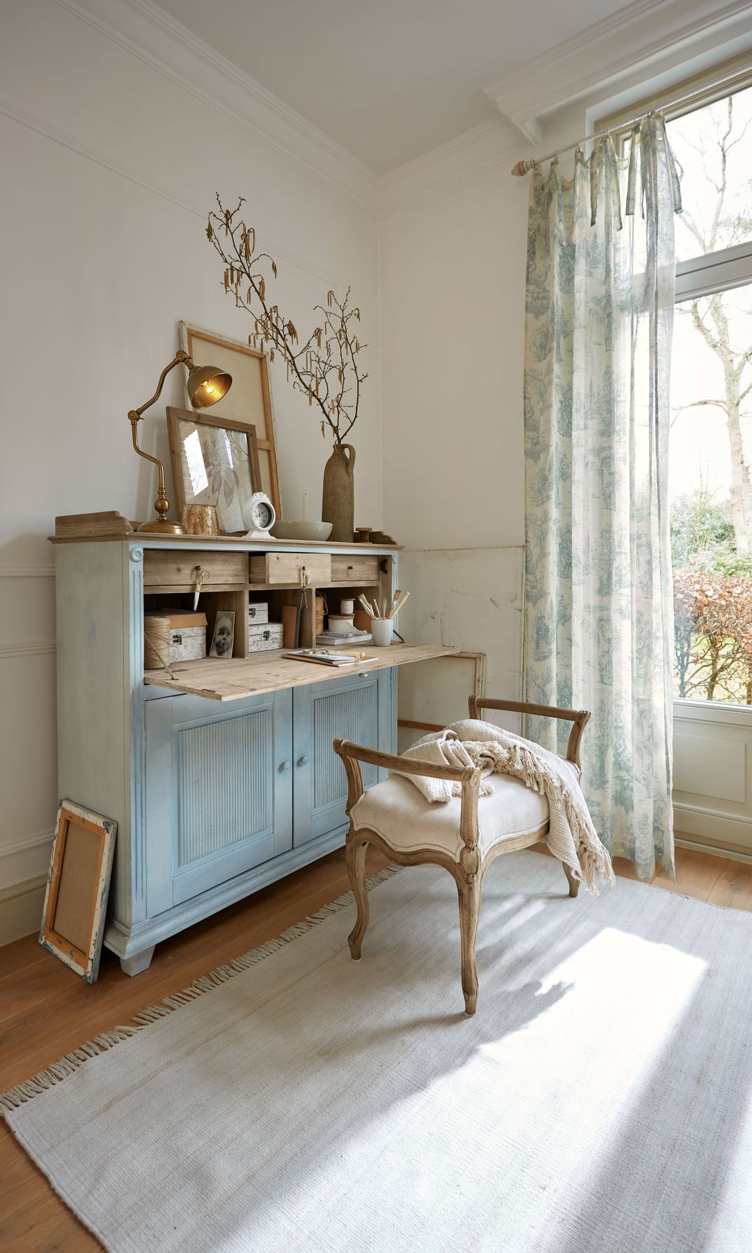 Secreter con tapa, banqueta tapizada, alfombra, jarrón, lámpara de sobremesa, cortinas ligeras de flores con lazos