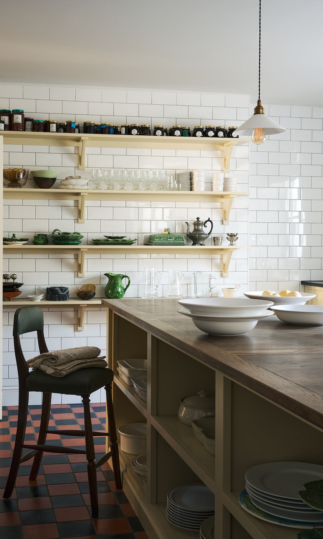 Cocina con isla, suelo de damero rojo y negro, isla abierta con vajilla, taburete, puerta de paso blanca y de cristal, baldas, lámpara de techo antigua