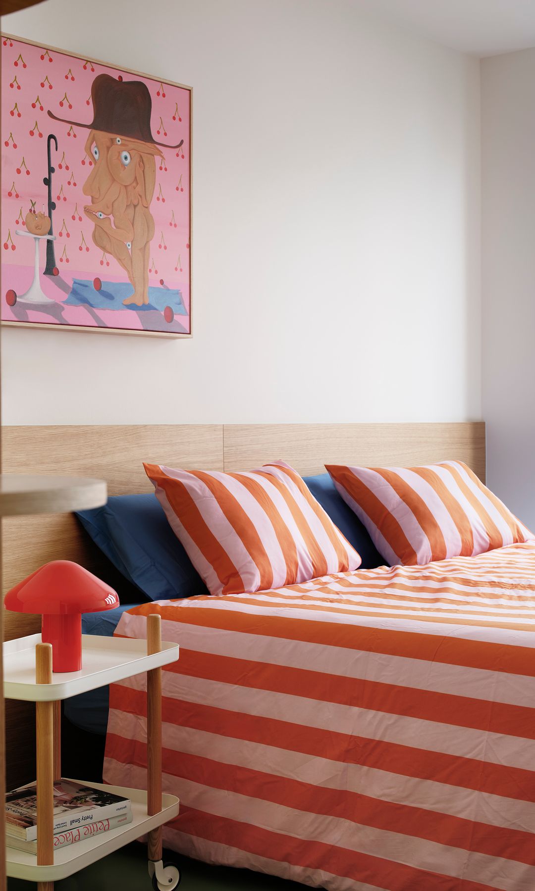 Dormitorio con funda de rayas naranjas y rosas, cuadro en la pared del cabecero, mesita de noche con ruedas, cabecero de madera