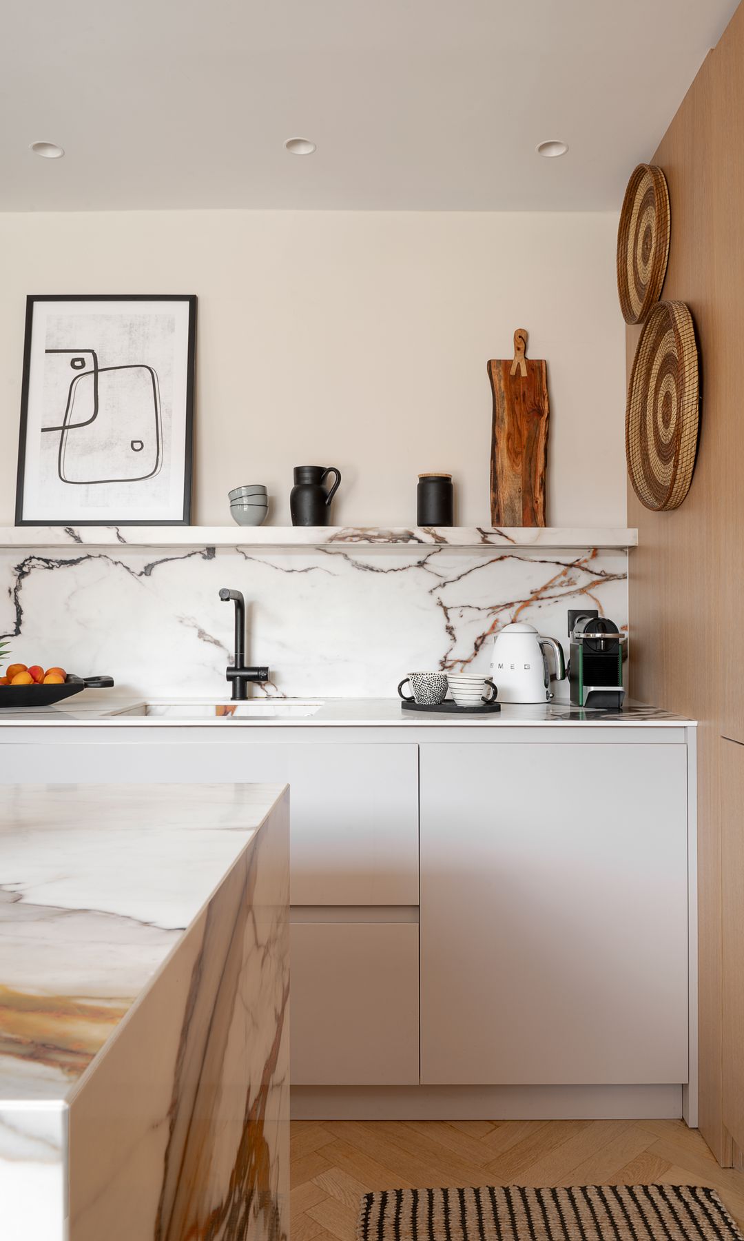 Cocina blanca con isla, sin armarios superiores, pared con un gran armario con frigorífico integrado, alfombra,bra, balda y antepecho de cerámica, con aspecto de mármol