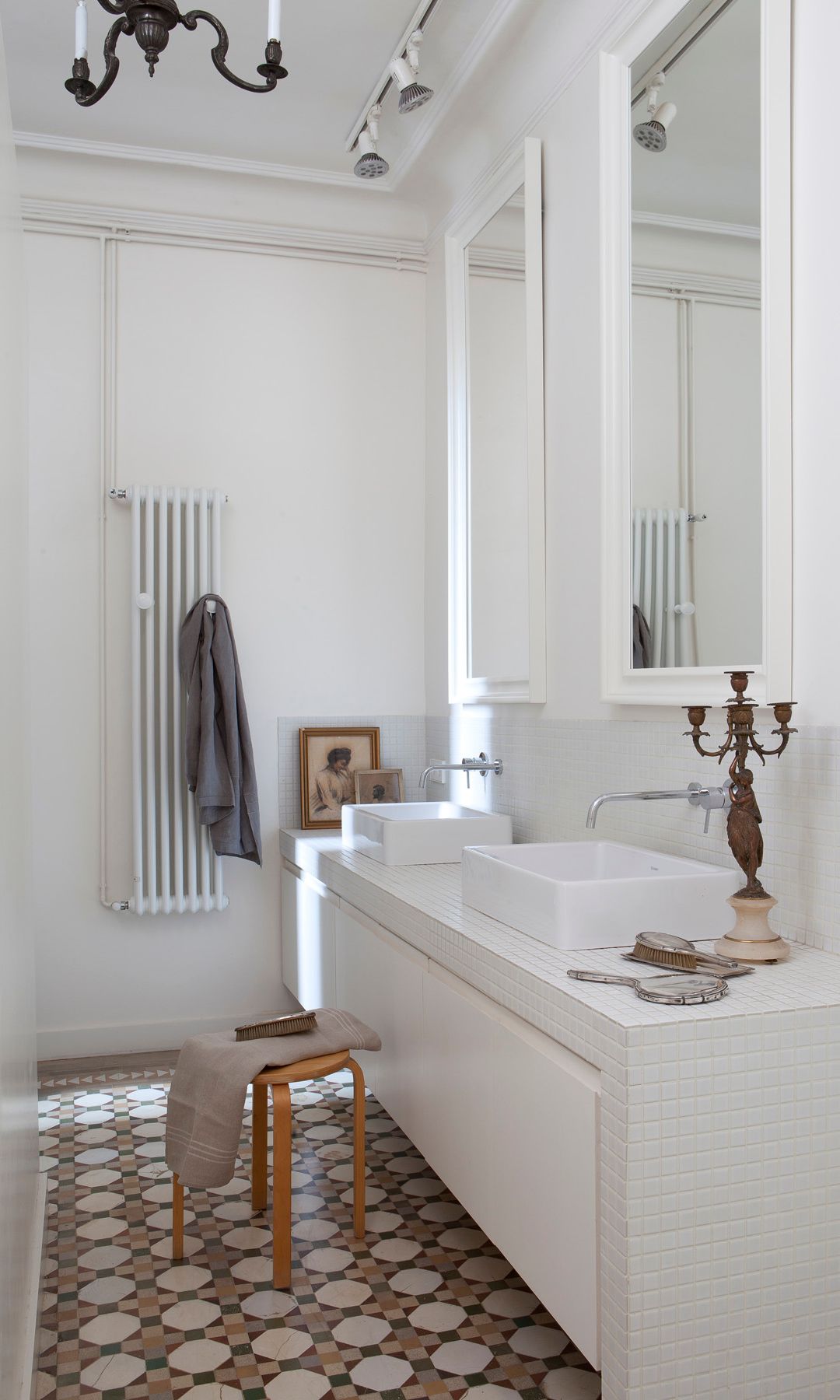 Baño con suelo hidráulico, espejos y muebles blancos, lámpara de araña, taburete de madera, cuadros antiguos, radiador toallero 