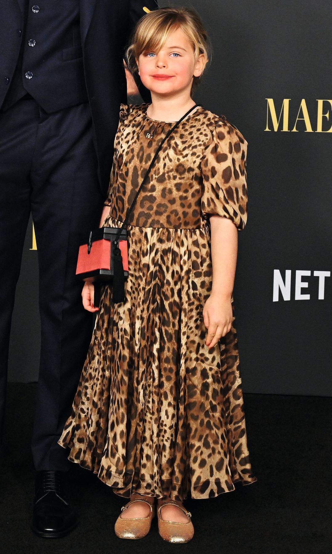 Christian Bale, en traje, posa con una niña en vestido leopardo en una alfombra roja.