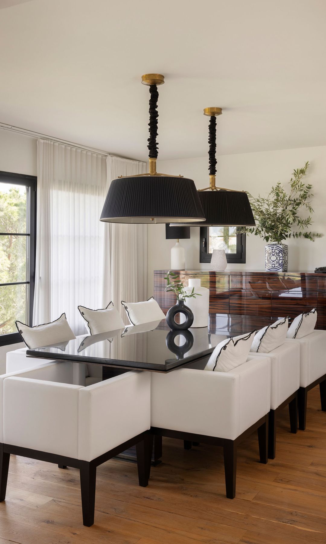Comedor con mesa rectangular negra, sillas tapizadas blancas, lámparas de techo negras, aparador de madera oscura, jarrón