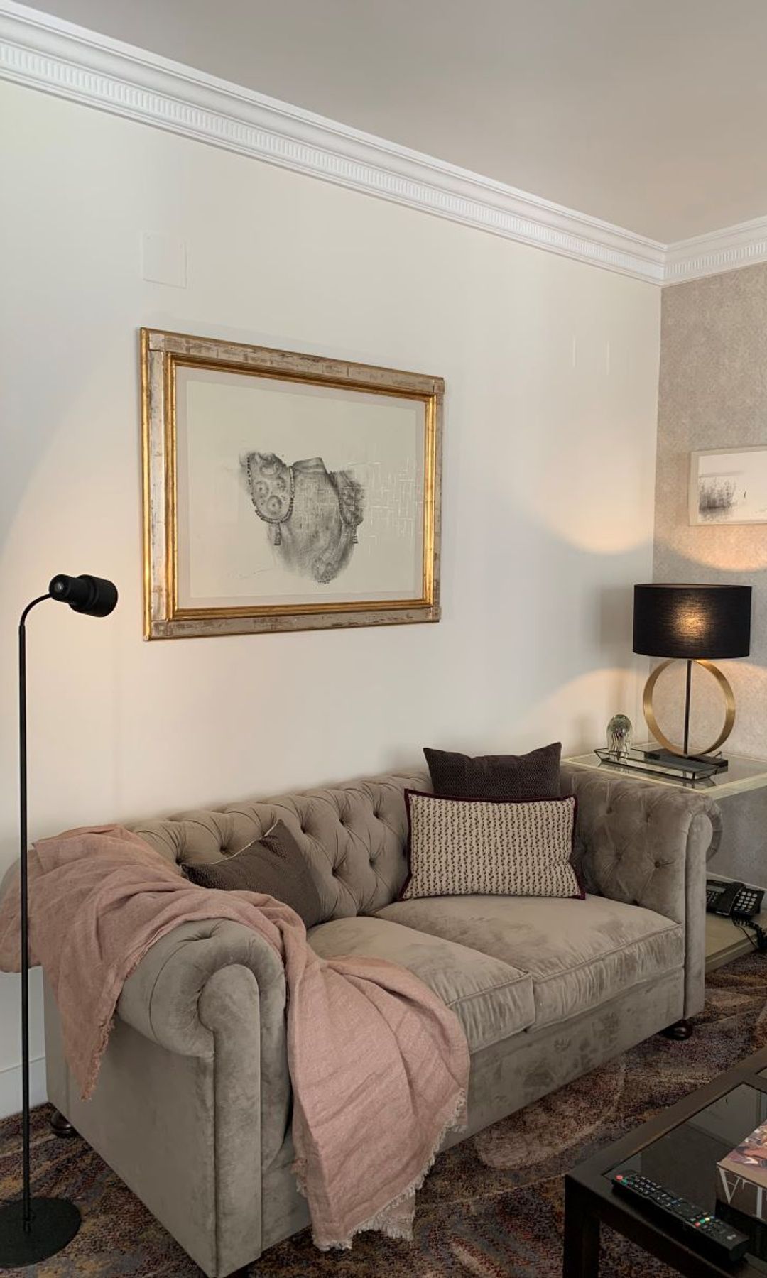 Salón con sofá gris con capitoné, molduras en el techo, cuadro con el marco dorado, manta srosa sobre el sofá, lámpara de pie negra, lámpara de sobremesa negra 